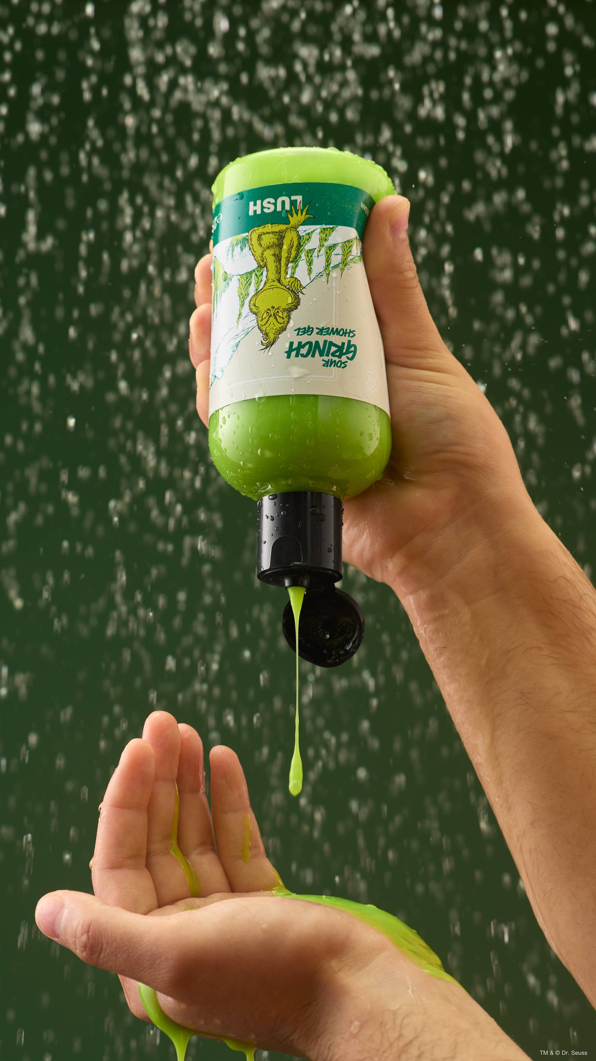 A hand holds an upside-down bottle of Lush 'Sour Grinch' bright green shower gel, squeezing a stream into an open palm amidst splashing water against a green background.  