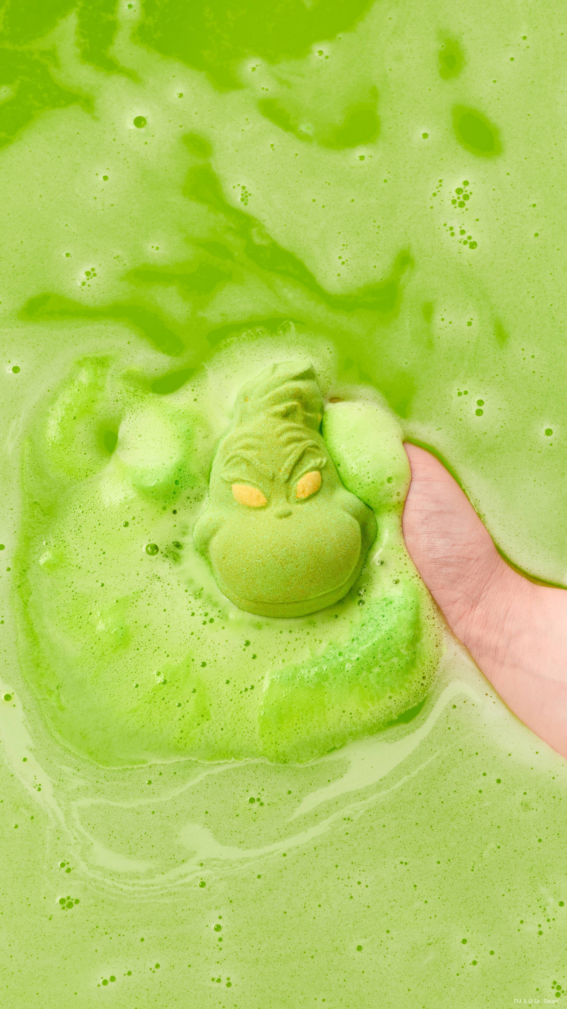 A hand holds a mischievous lime-green "Grinch" face-shaped Lush bath bomb in bathwater as it dissolves, creating a sea of bright green water surrounded by thick, vibrant green foam.  