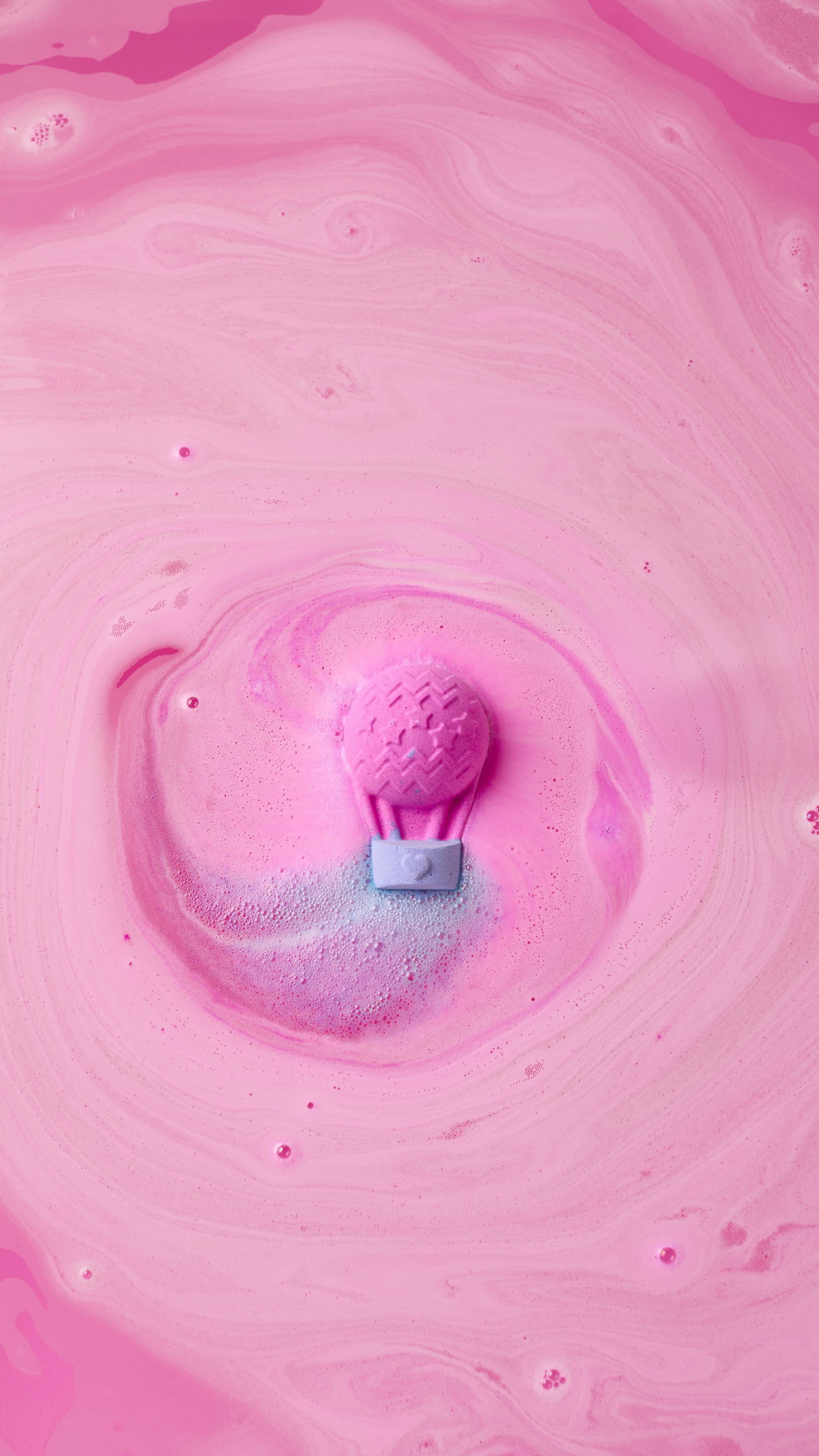 A close-up, aerial view of a pink "Up, Up And Away" hot air balloon-shaped bath bomb dissolving in water. It creates a swirling pattern of vibrant pink and soft lilac foam that spirals across the surface of the deep pink bathwater.