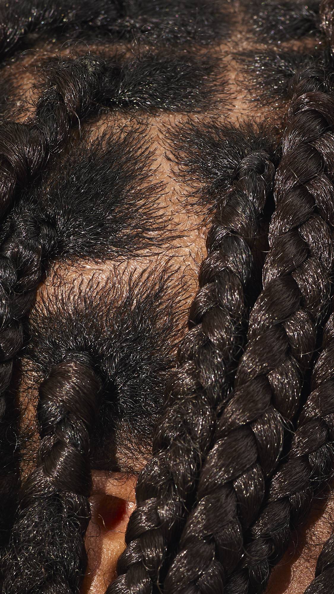 An extreme close-up of a person's scalp showing the intricate parting and base of thick, dark, shiny braids. The hair is neatly sectioned into a grid pattern, with the tightly woven braids extending downwards.