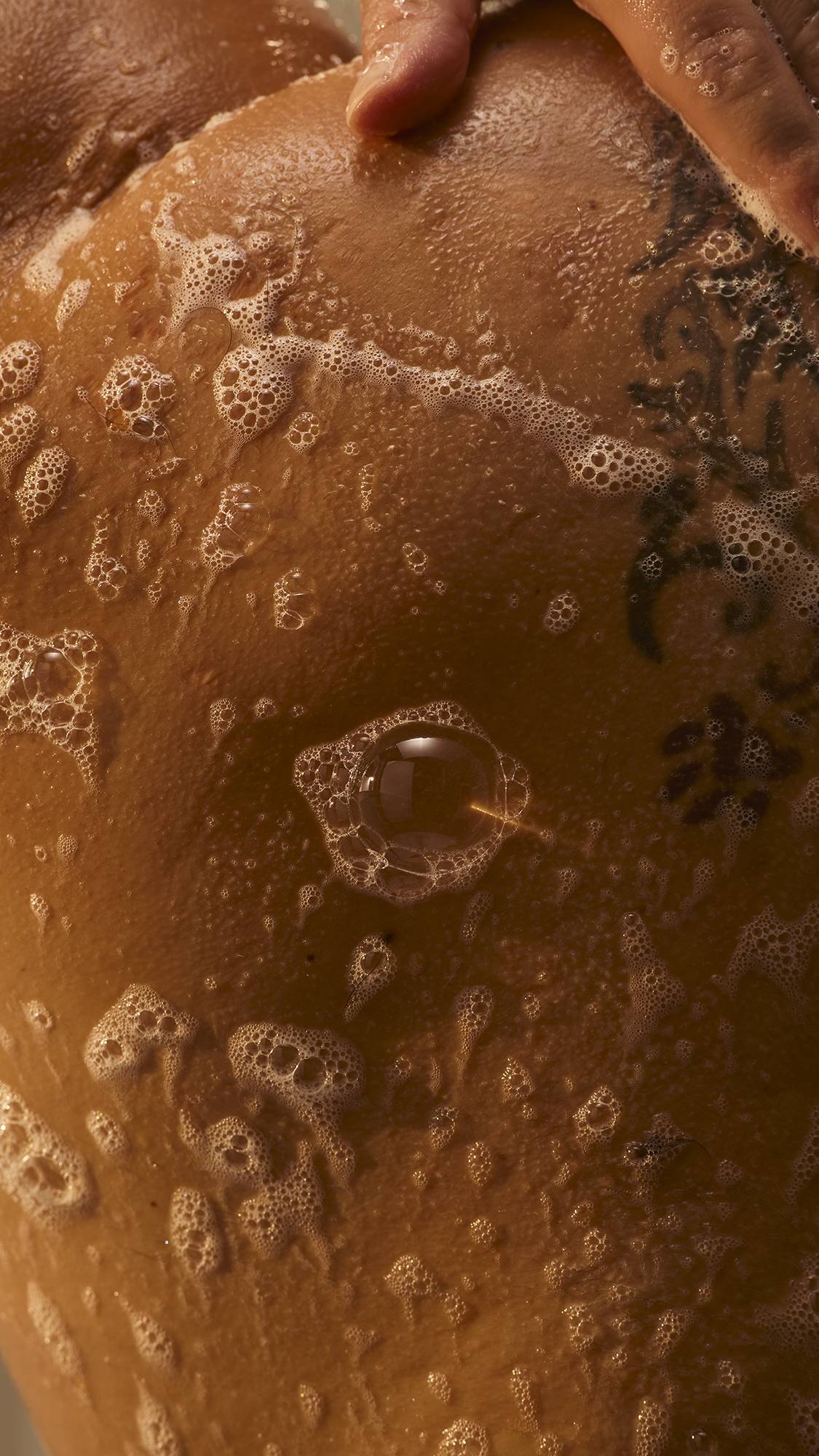 Close-up of soapy white suds and delicate bubbles spread across tattooed, wet skin as a hand washes in warm lighting.