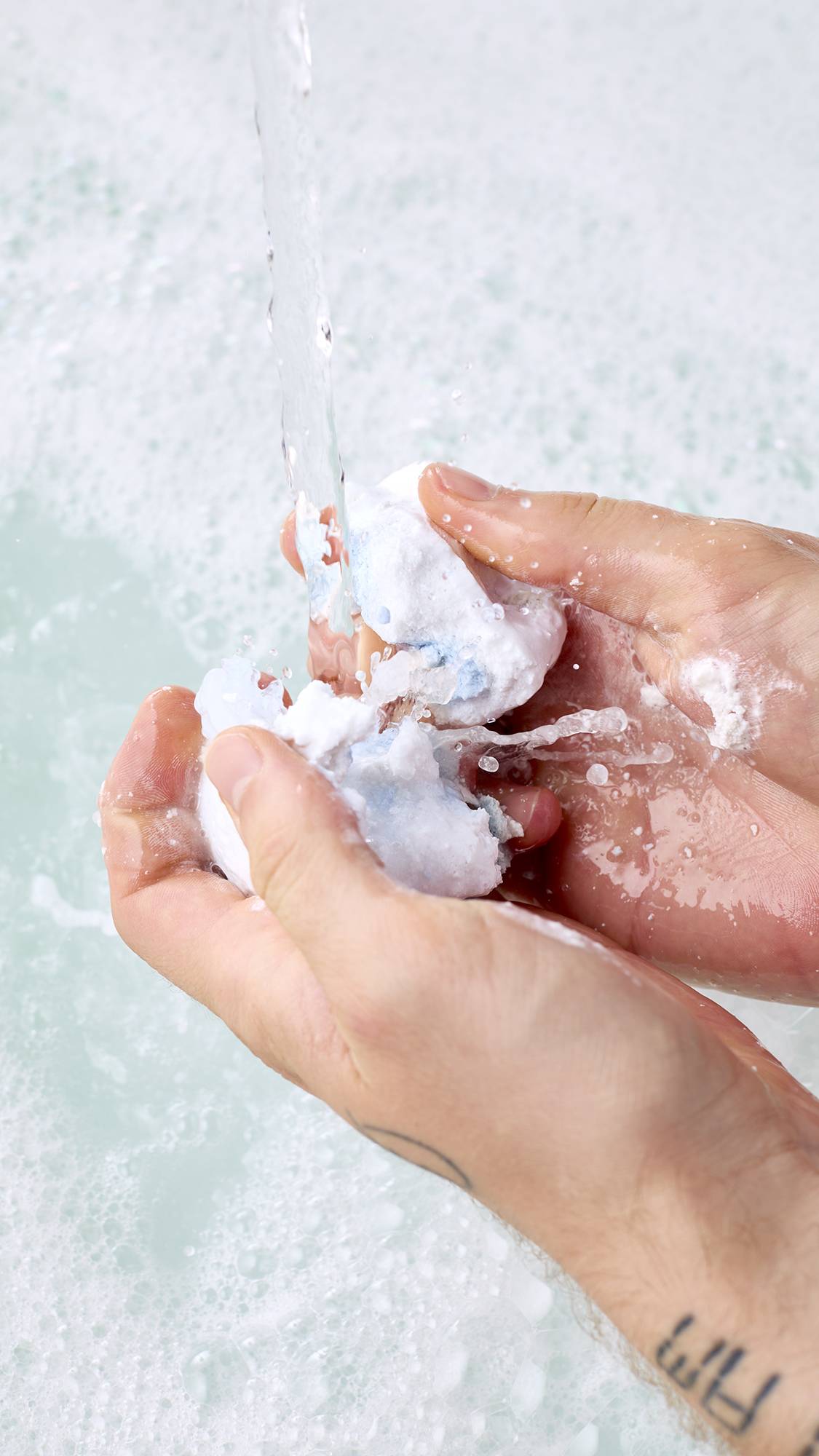 Two hands crumble a white and pale blue sleeping fox bubble bar under running tap water, splashing over milky white bathwater filled with light, frothy bubbles.
