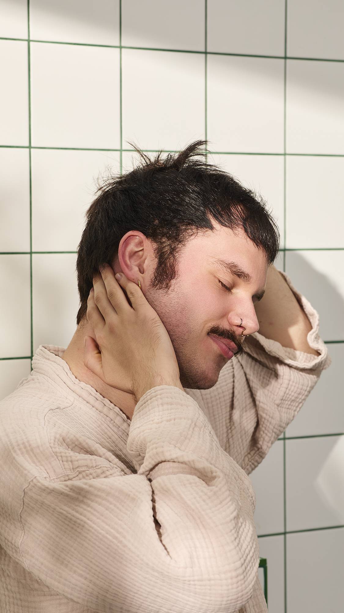 A person with short dark hair and a moustache smiles with their eyes closed, wearing a light pink robe. They are resting their hands on their clean hair against a background of white square tiles with green grout.
