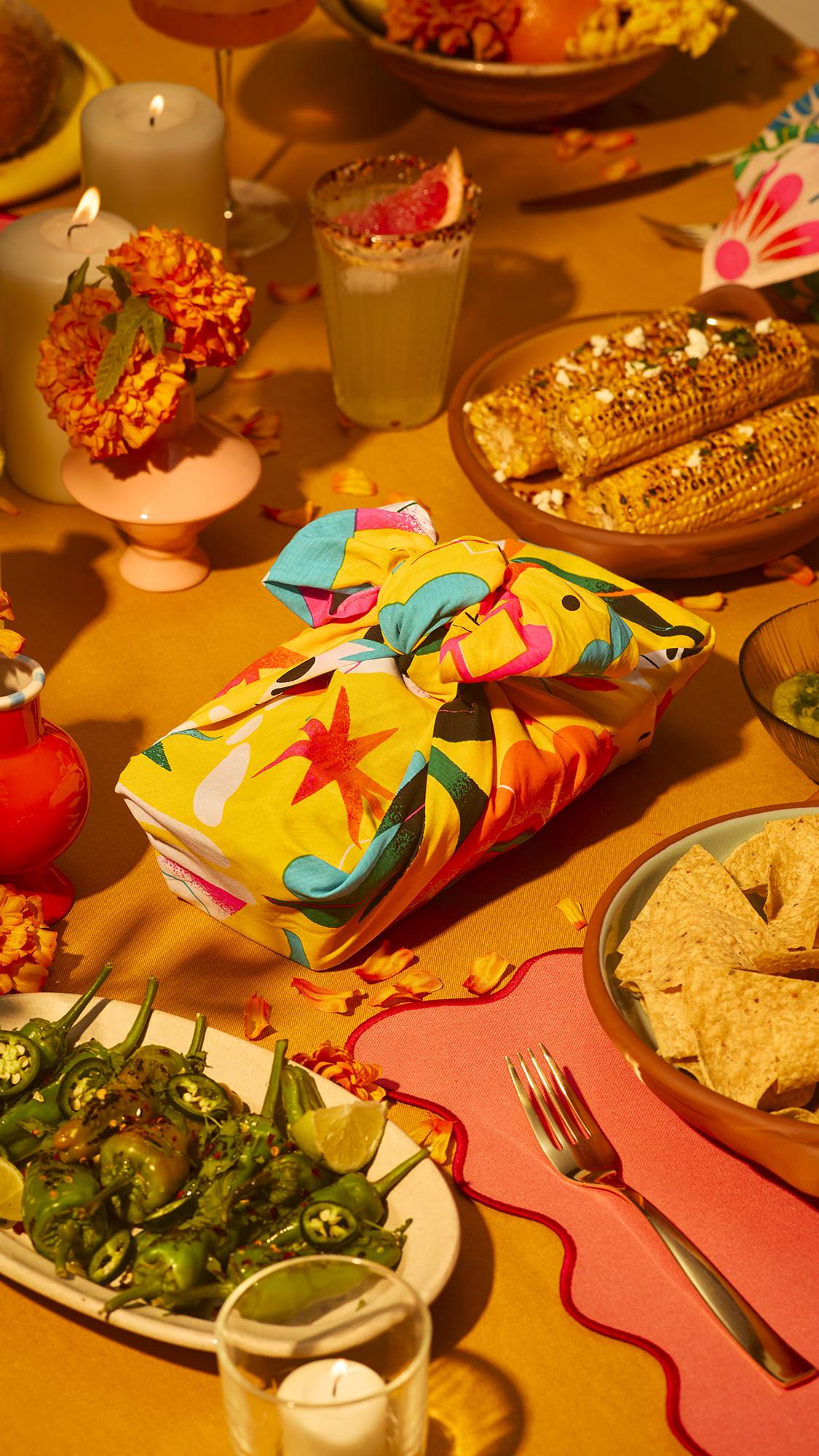 A vibrant yellow knot-wrapped gift box sits at the centre of a festive dining table surrounded by Mexican dishes like grilled corn, tortilla chips, and green peppers, lit by soft candles and vibrant marigold flowers.