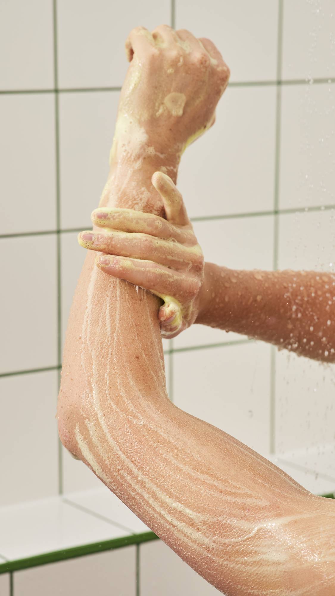A person washes their arm in the shower, smoothing soft yellowish-white soapy lather up their wet forearm under running water against a white tiled wall.