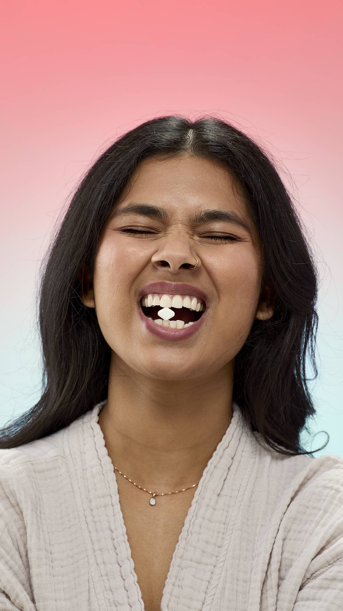 A person with long dark hair and a nose ring smiles with their eyes closed, holding a small, diamond-shaped Banana Bread mouthwash tab between their front teeth.