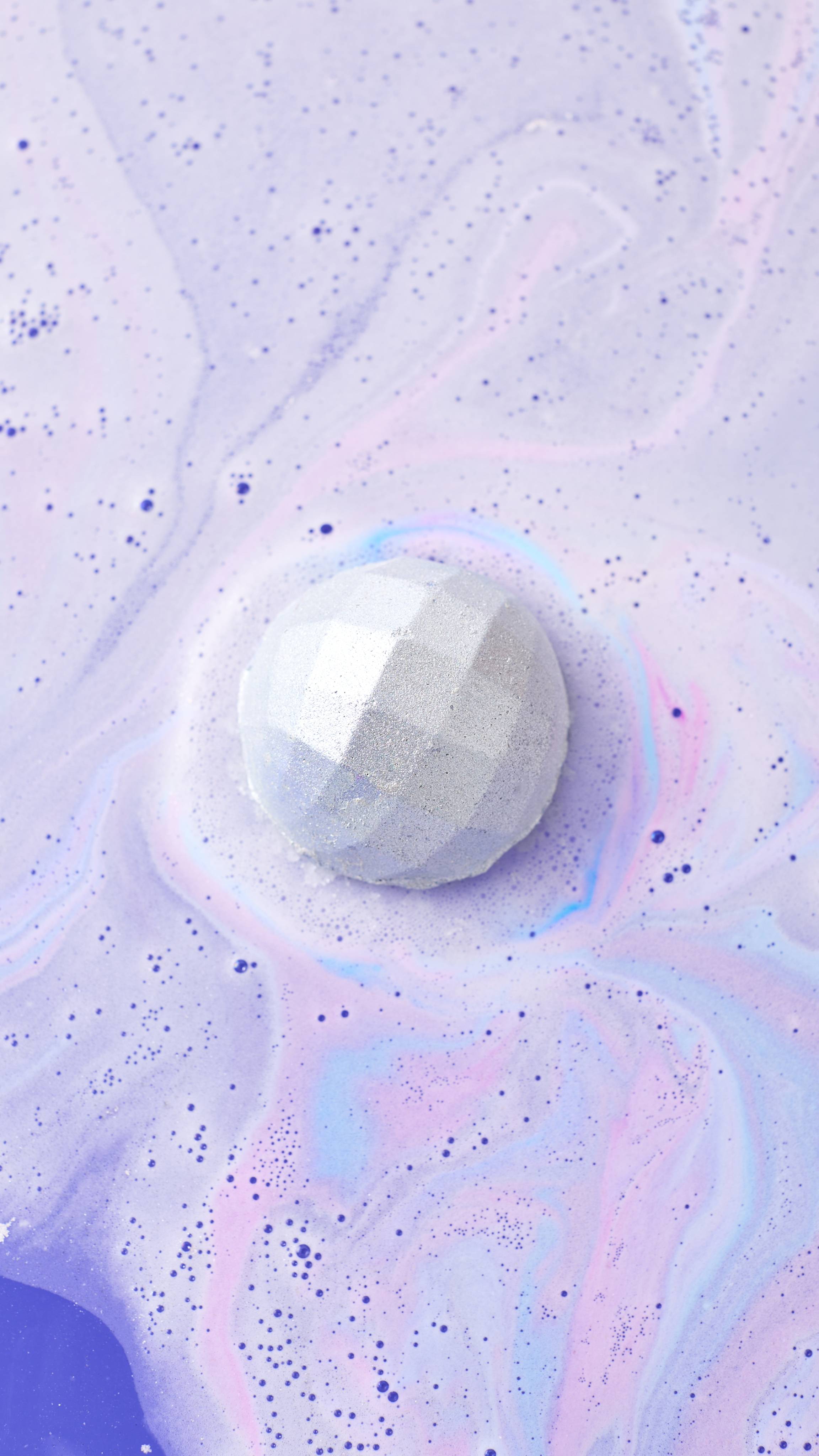 A shimmering, lilac "Bathe All The Time" bath bomb with a faceted, diamond-like surface dissolves in water. It releases swirling clouds of pale purple, pink, and blue fizz, creating a dreamy, marbled pattern across the surface of the bathwater.