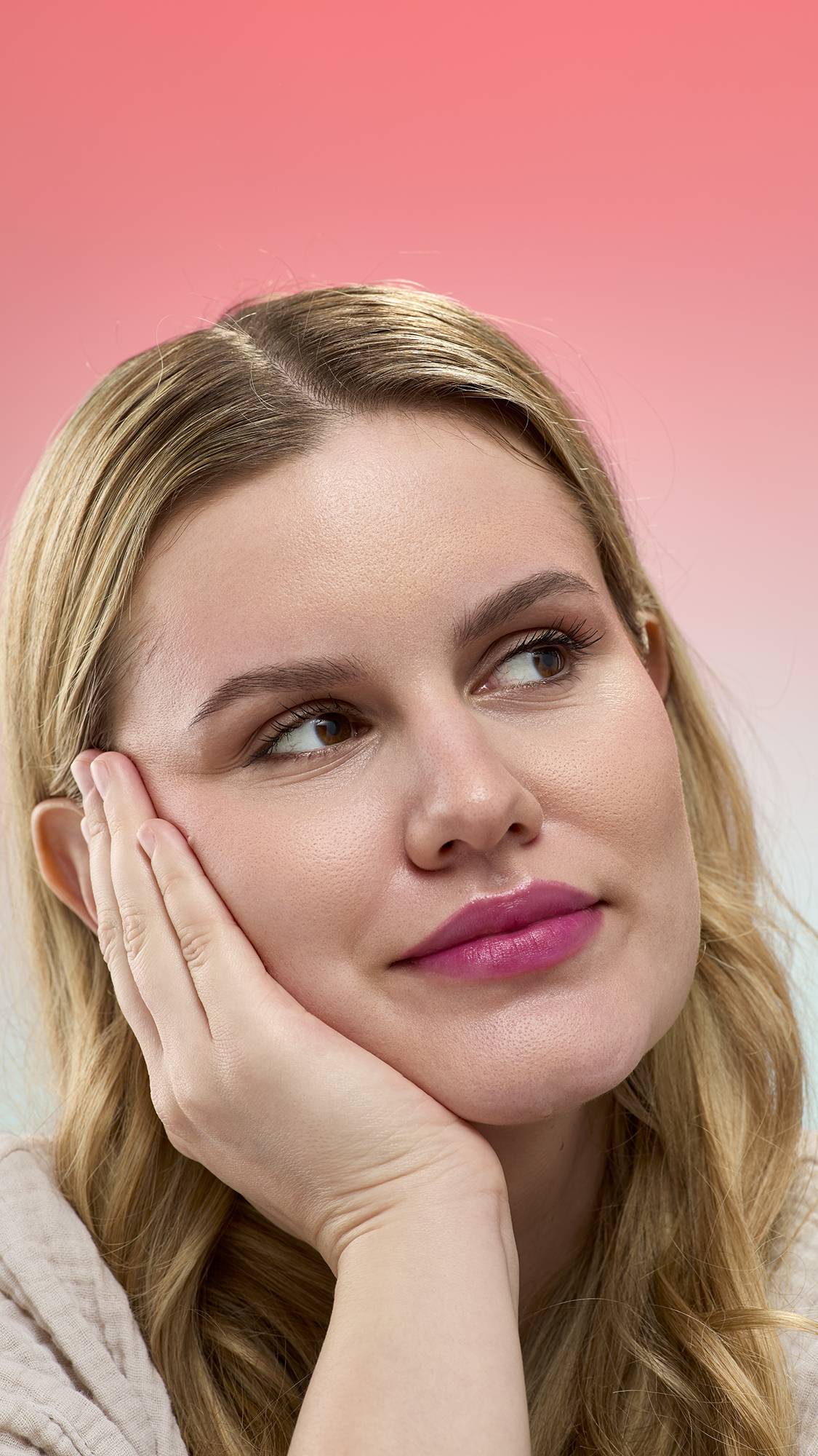 A person with blonde wavy hair rests their face on their hand, looking away while wearing a deep berry-colored Black Rose Lip Stain. They are set against a pink gradient background and wearing a light, textured robe.