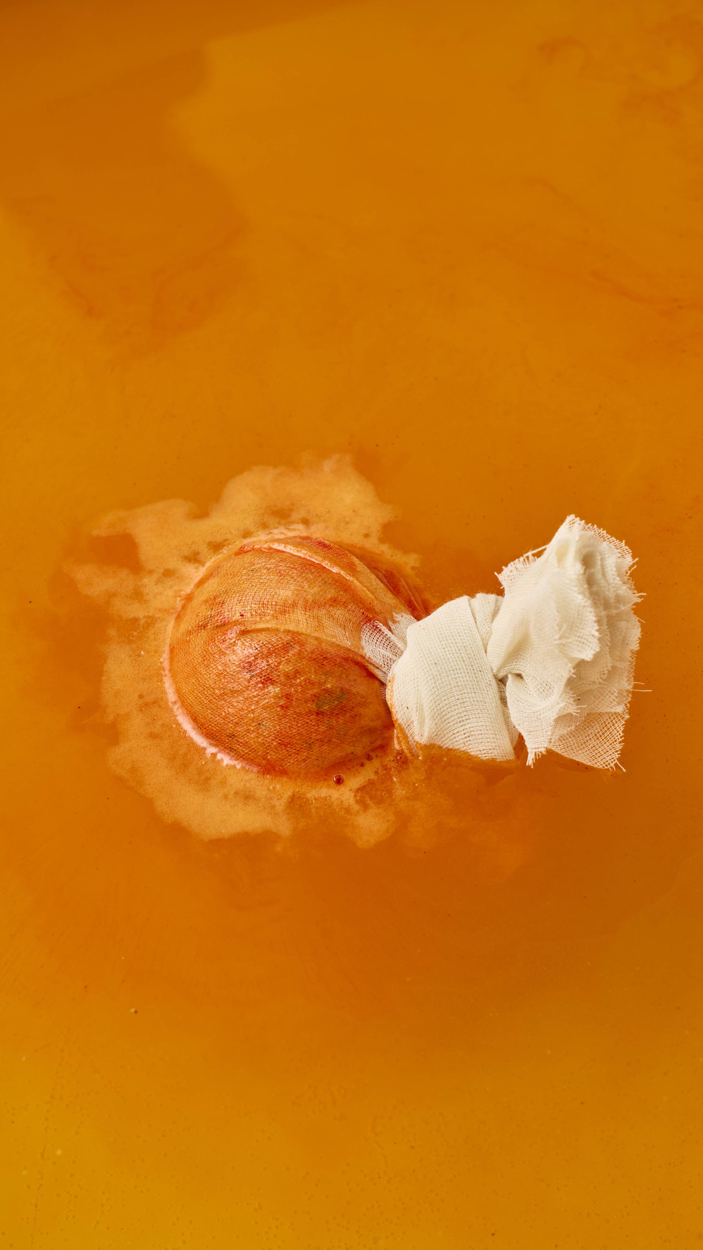 A round bath bomb, wrapped in a tied cheesecloth pouch, floats and fizzes on top of vibrant orange bathwater. White foam creates a soft ring around the fabric pouch as it dissolves.