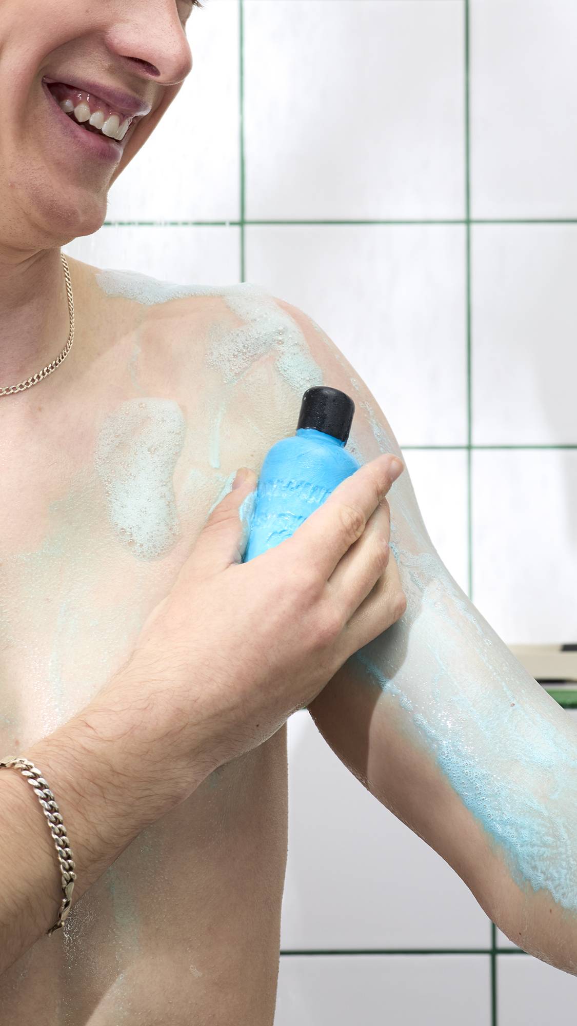 A side-profile shot of a smiling person lathering their arm in the shower with a bright blue, bottle-shaped solid shower gel. A light blue, frothy lather coats the skin against a background of white tiles with green grouting.