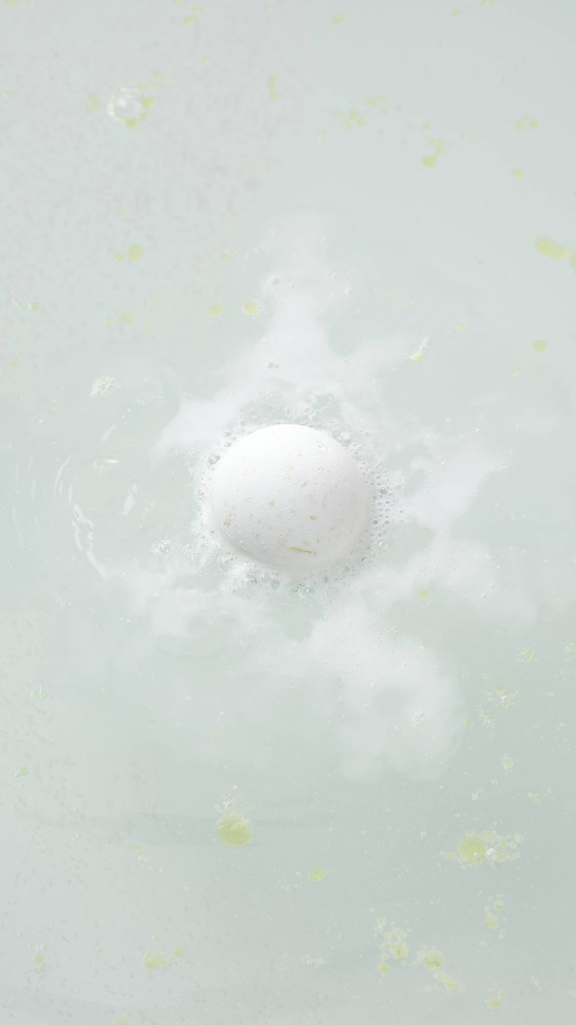 The white, spherical Butterball bath bomb sits on a plain, light gray background. The product features a matte, slightly textured surface with small, visible brown flecks of cocoa butter throughout.