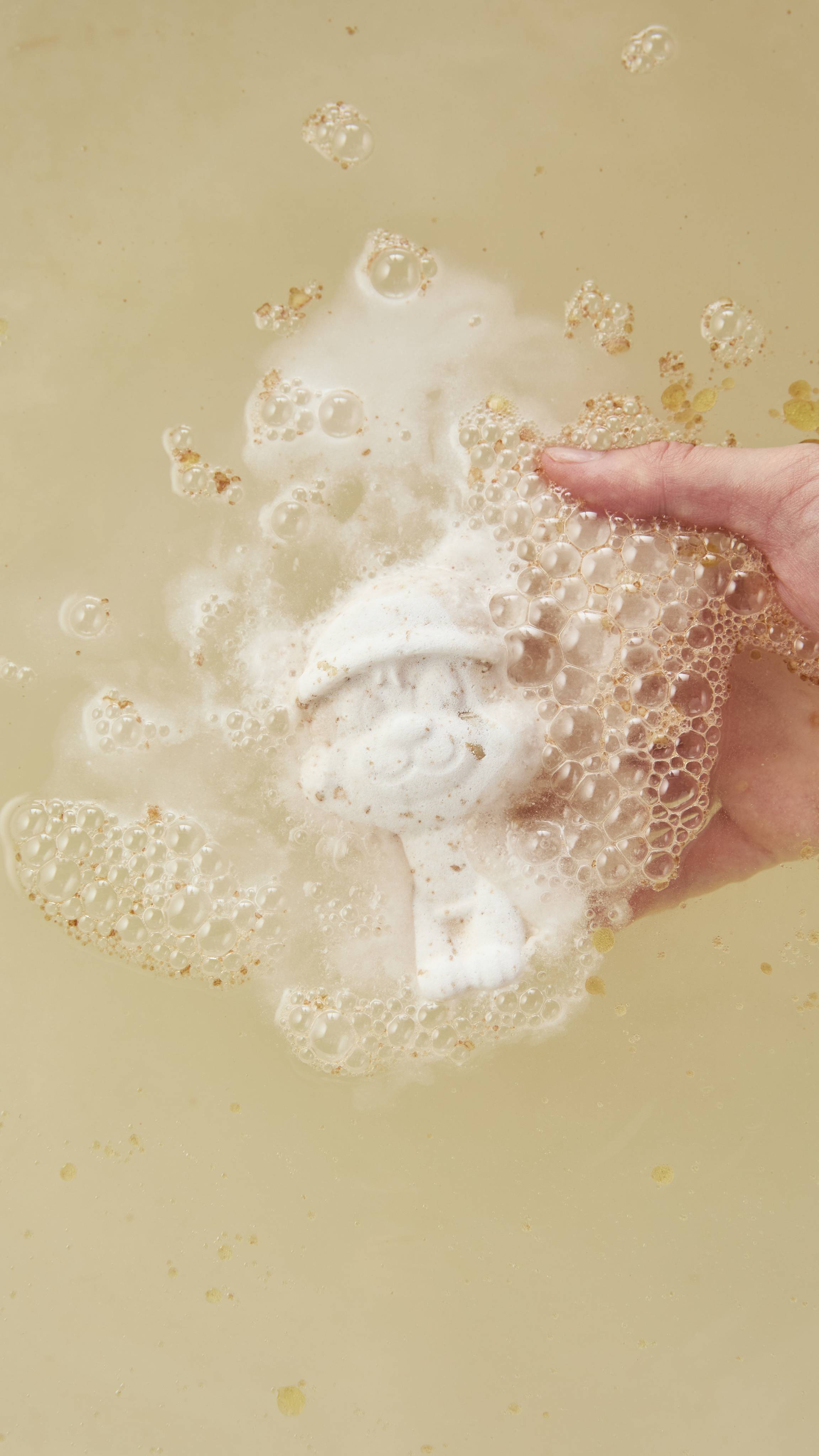 A white polar bear bath bomb dissolves under milky bathwater, producing a froth of bubbly white foam as a hand holds it beneath the surface.