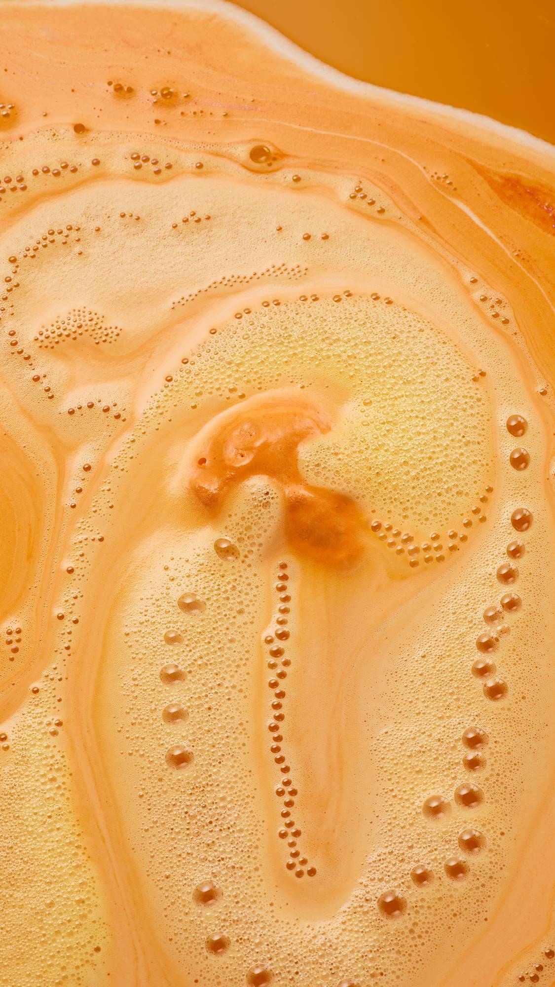 Overhead close-up of vivid orange bathwater with swirling patterns of creamy, pale orange foam and delicate lines of fizzing bubbles across the surface.