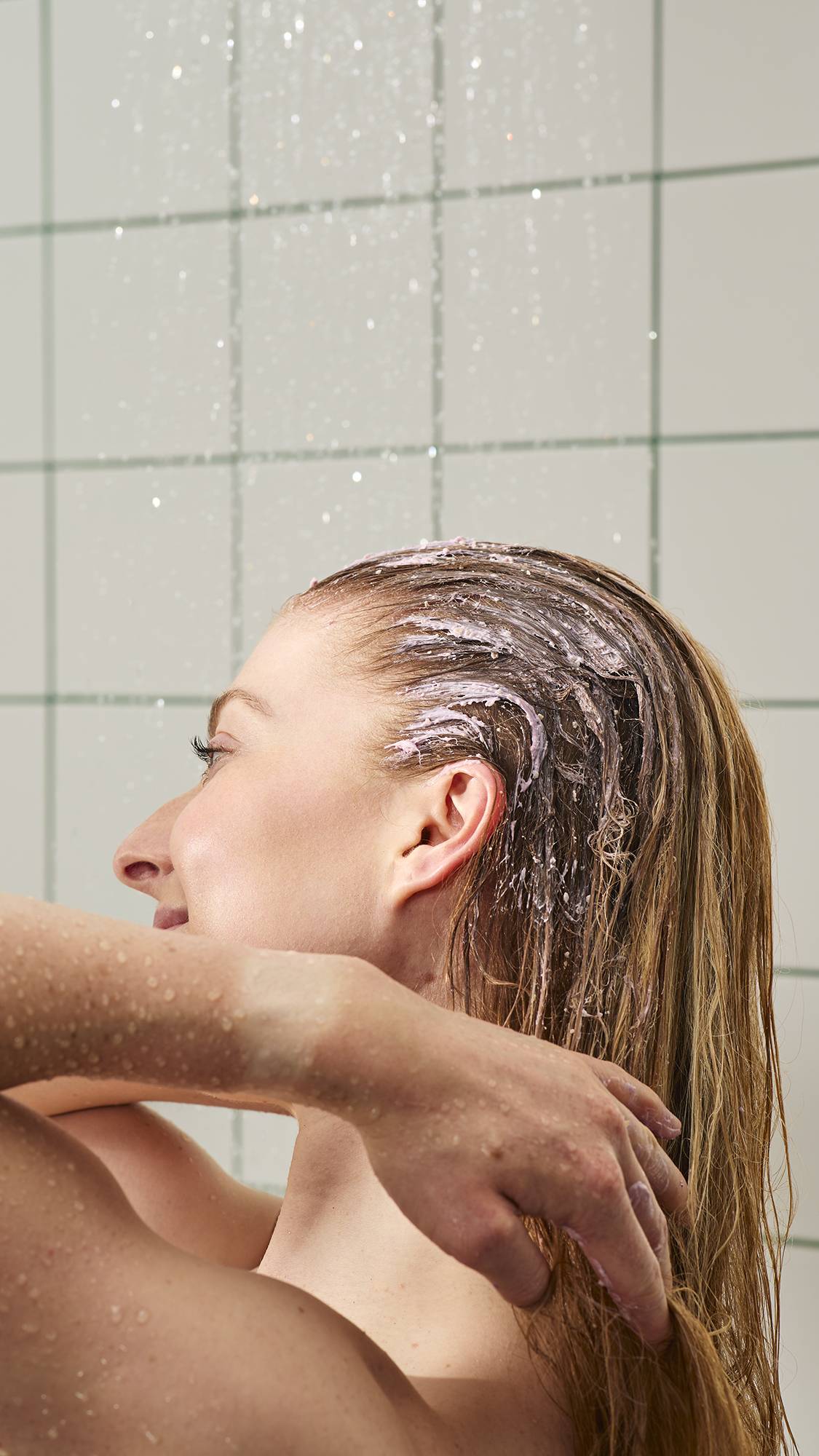 A close-up shows a person in a shower, massaging the "Calm Down" lilac-coloured treatment into their long, wet reddish-brown hair. Water droplets spray in the background against white square tiles with green grout. 