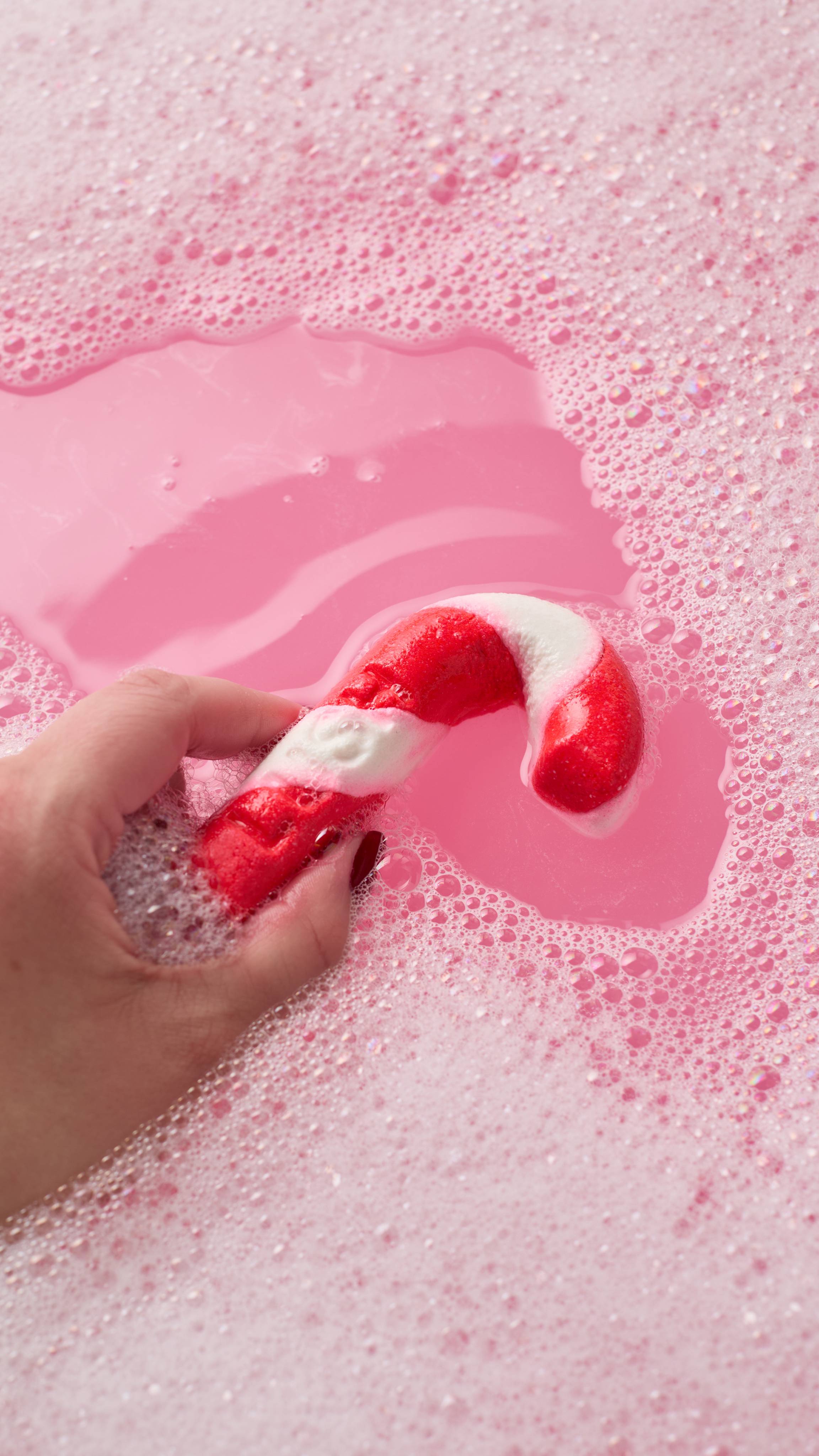 A hand swishes a red and white candy cane bubble bar through vibrant pink bathwater, parting a thick layer of fine white bubbles and foam.