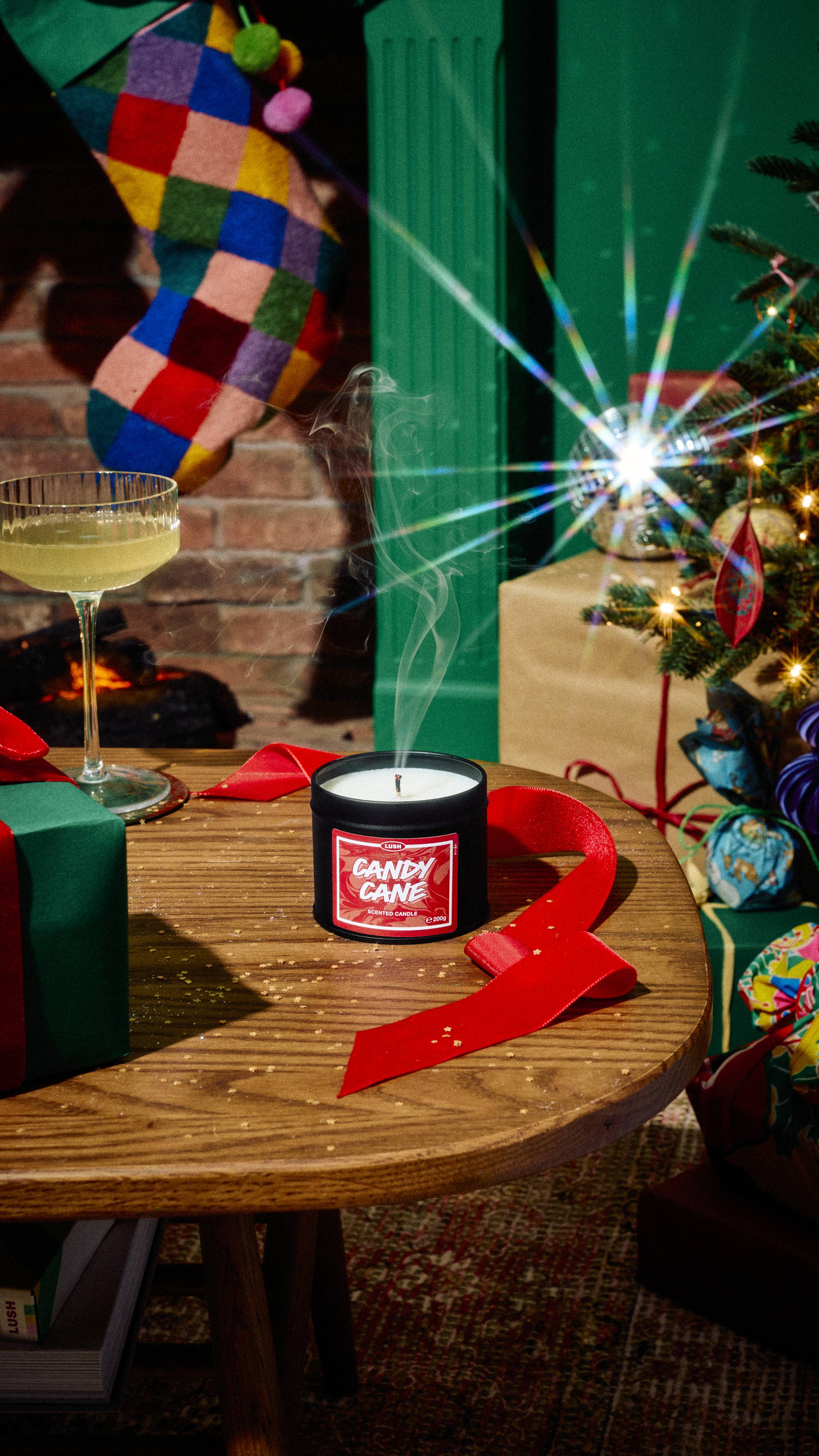 Smoke rises from a freshly extinguished tin of Lush Candy Cane scented candle on a wooden table, beside red ribbon, a cocktail glass, and a gift, set in a festive room with a Christmas tree and colourful stocking.