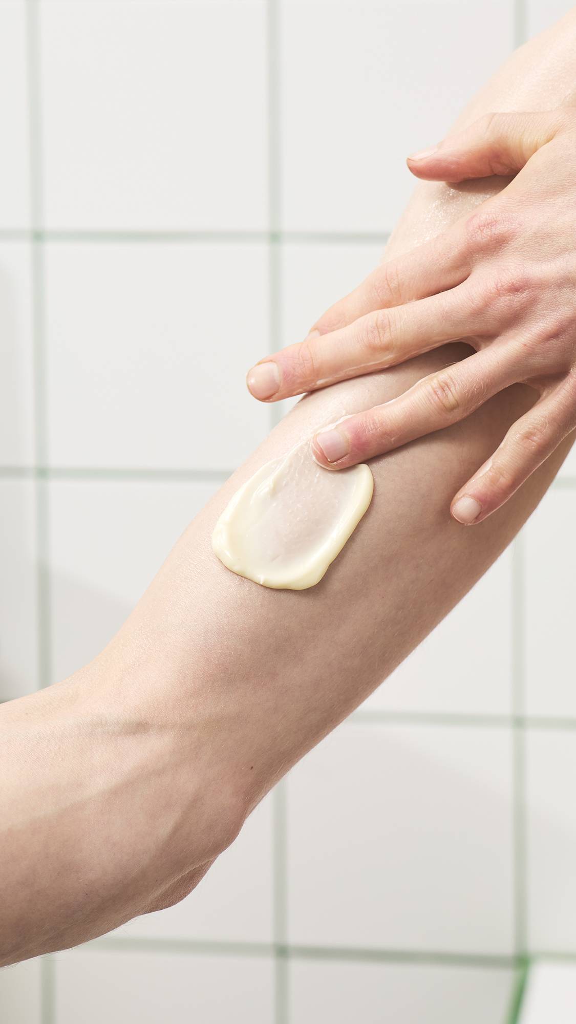 A hand gently smooths a swirl of creamy, off-white body lotion onto a forearm against a white tiled bathroom wall background.