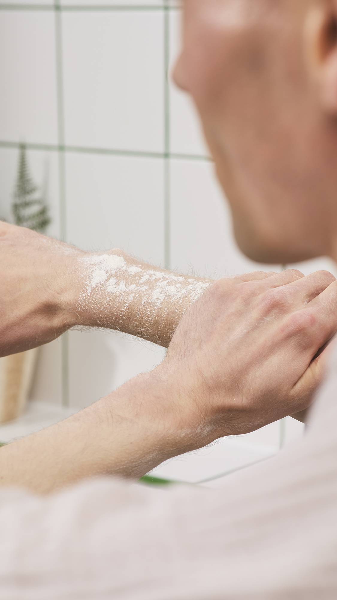 A person smooths fine white dusting powder across their forearm using their opposite hand against a white tiled bathroom background.