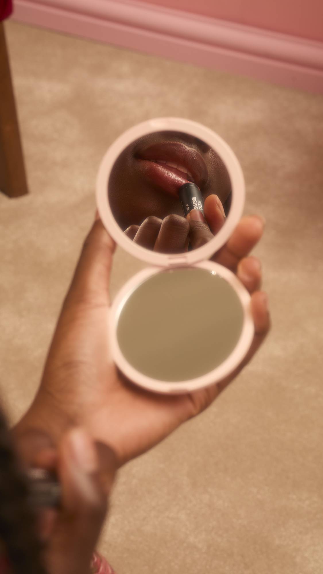 A person with dark skin holds a small, round, pink compact mirror, reflecting their lips as they apply the Lush "Cherry Bubblegum" lip oil. The background shows a soft beige carpet and a hint of a pink wall.