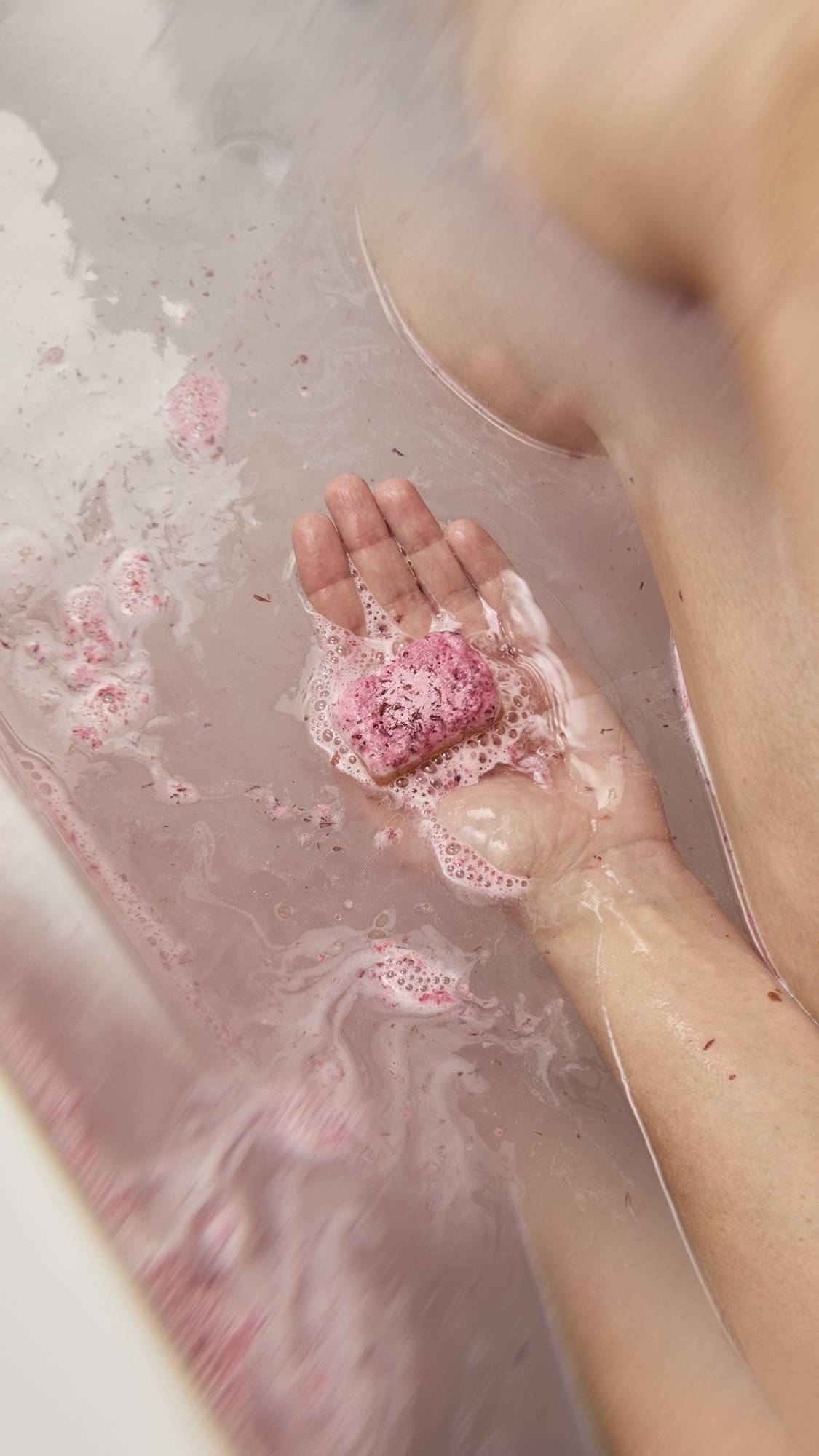 A hand holds a pink, textured "Cherry Pie" scrub bar underwater. The bathwater is milky pink with floating red exfoliating bits and foam.