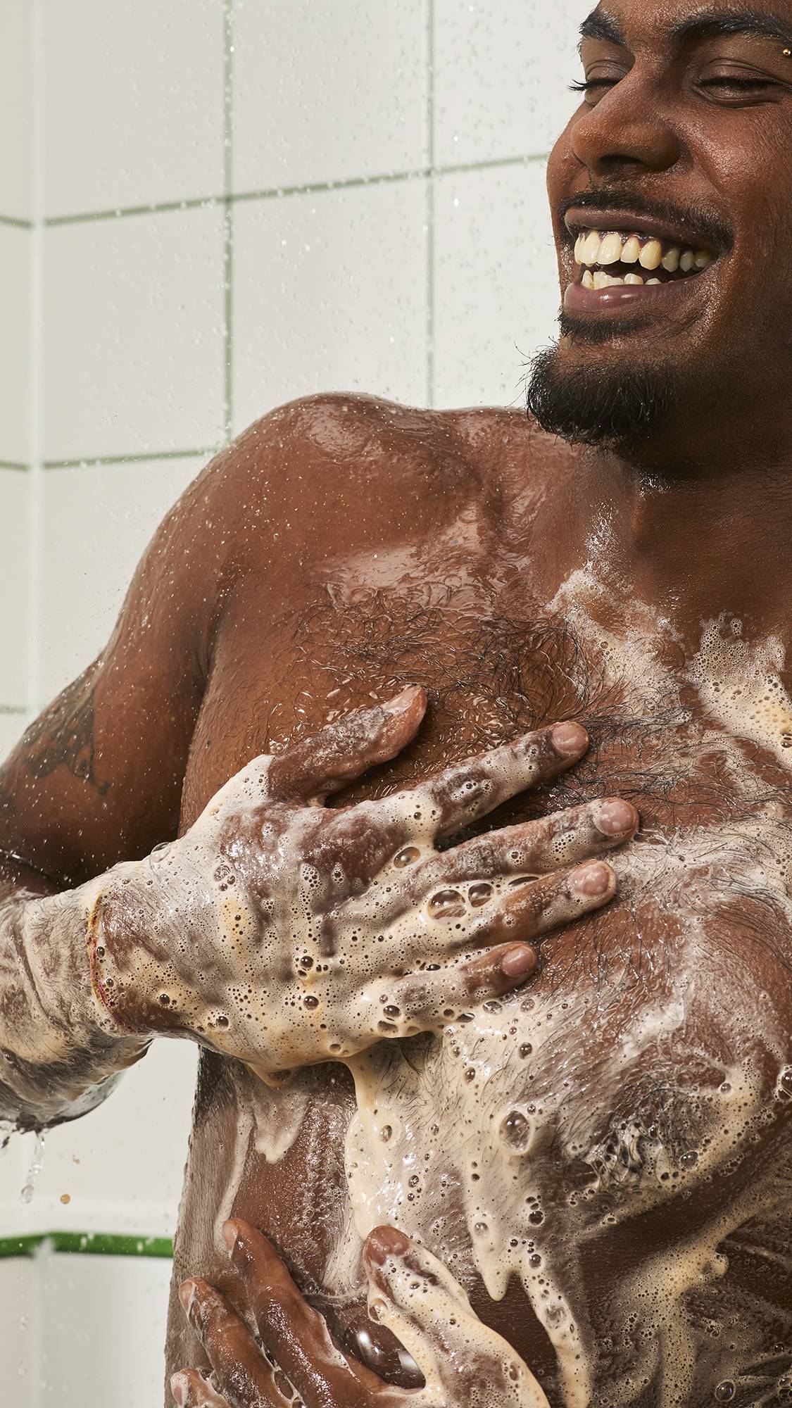 A smiling model with a beard lathers shower gel across their chest and torso, hand covered in rich white soapy suds against a white tiled shower wall.