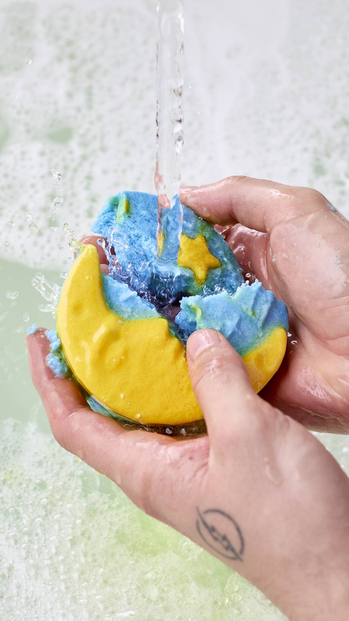 Two hands crumble a blue and yellow moon "Christmas Eve" bubble bar under running tap water, splashing over soft white bath foam.