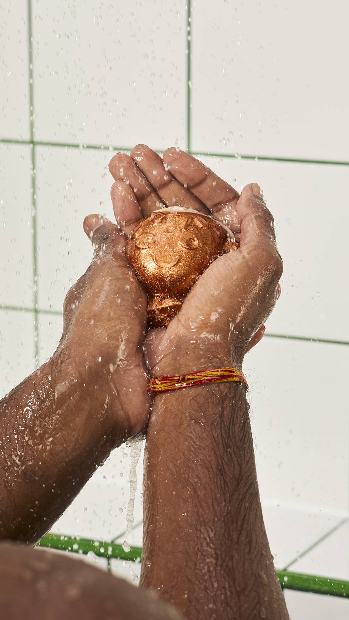 A pair of wet hands cradles a shimmering bronze Lush "Christmas Pud" character jelly under falling water in the shower, with a white tiled wall in the background.