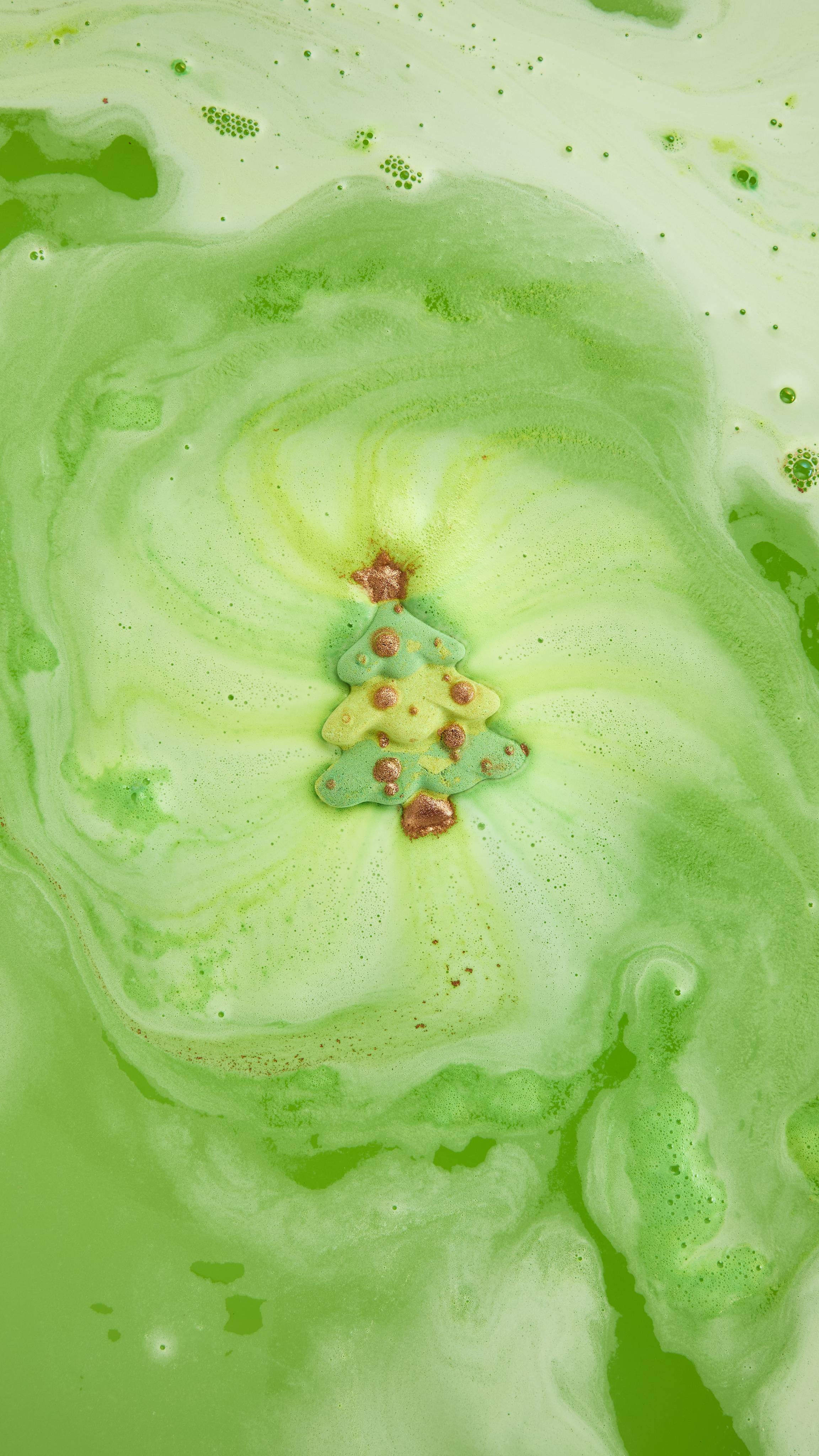 A green Christmas tree bath bomb with gold accents dissolves in bright green bathwater, creating a swirling vortex of shimmering, pale green foam.