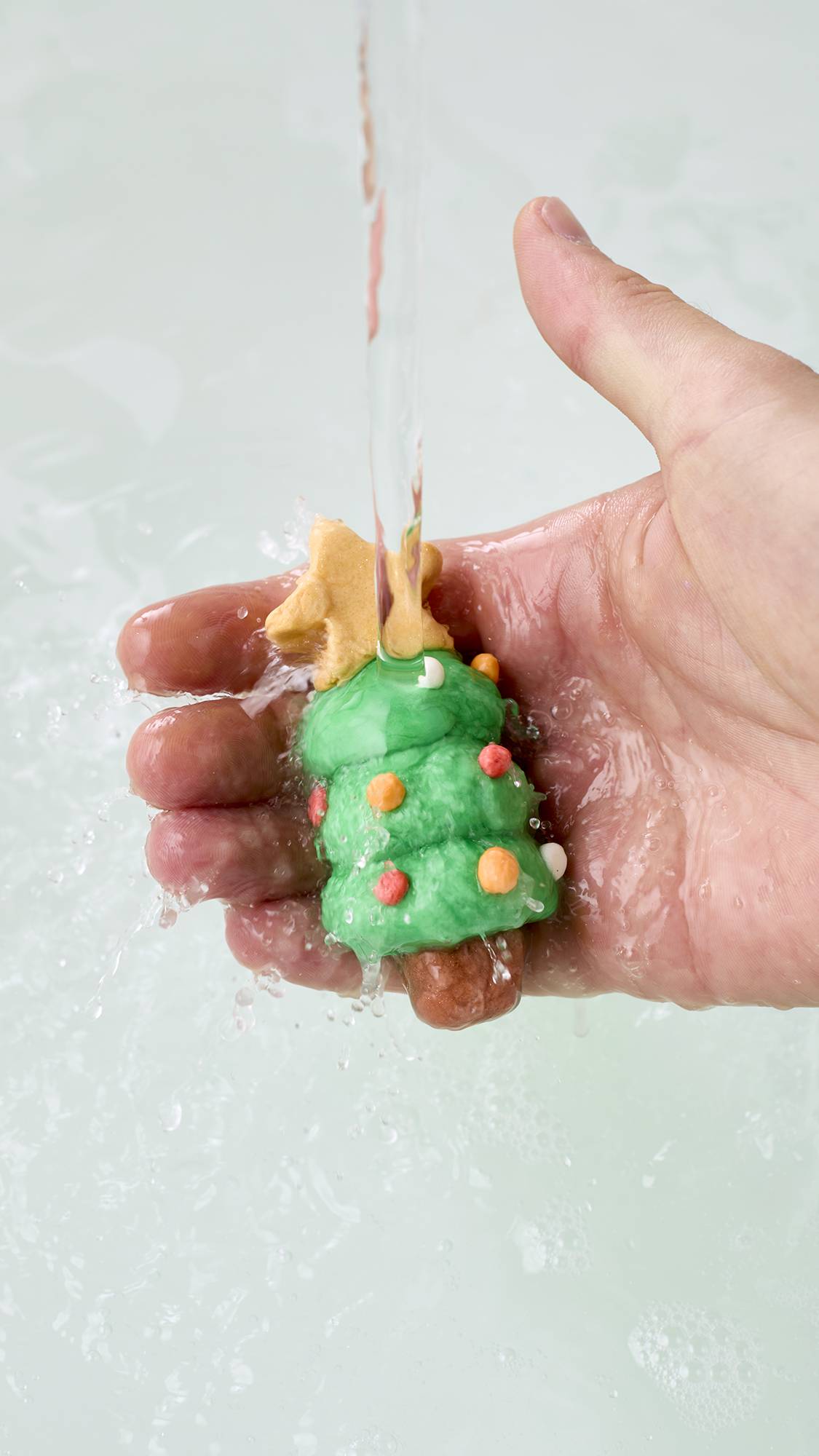 A hand holds a small green Christmas tree-shaped model, crafted from the Lush "Christmas Wreath" FUN product, topped with a yellow star and colourful baubles under running tap water over clear bathwater, splashing gently.