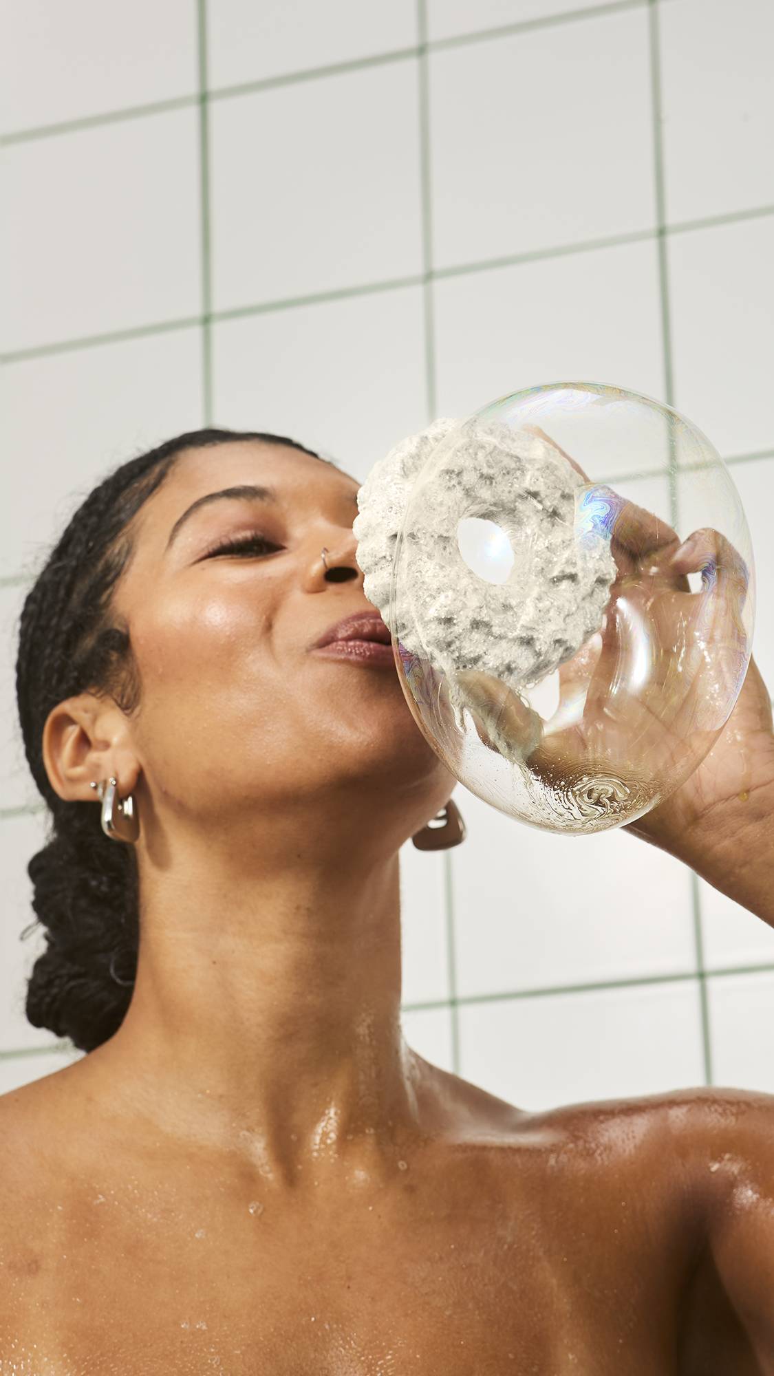 A smiling woman in the bath blows a large, iridescent soap bubble through the central hole of a white, wreath-shaped bubble bar held up to her mouth, set against a white tiled wall.