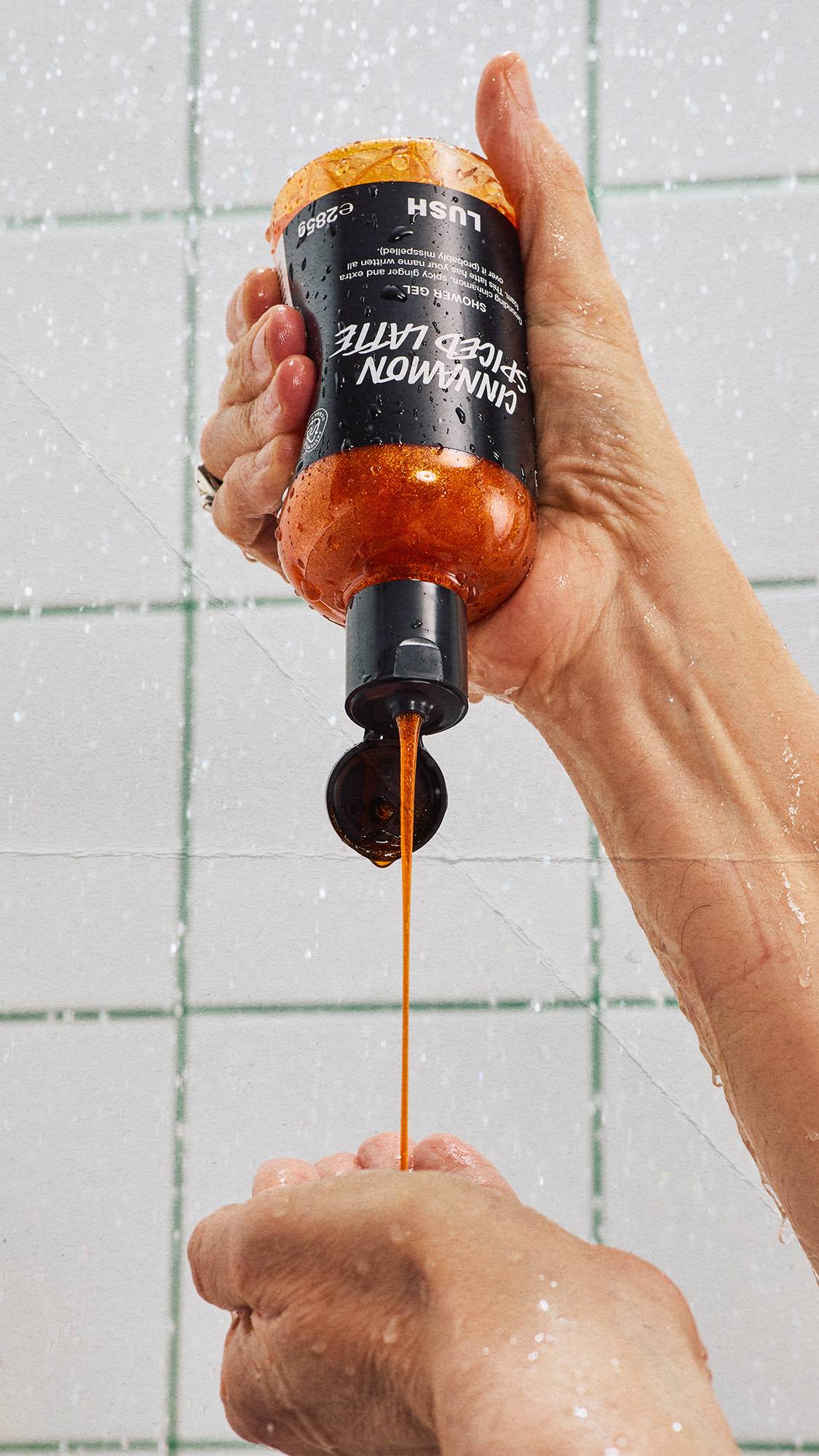 A hand holds an upside-down bottle of Lush Cinnamon Spiced Latte shower gel, pouring a steady stream of shimmering copper liquid into the other open hand against a backdrop of white tiles with green grout.
