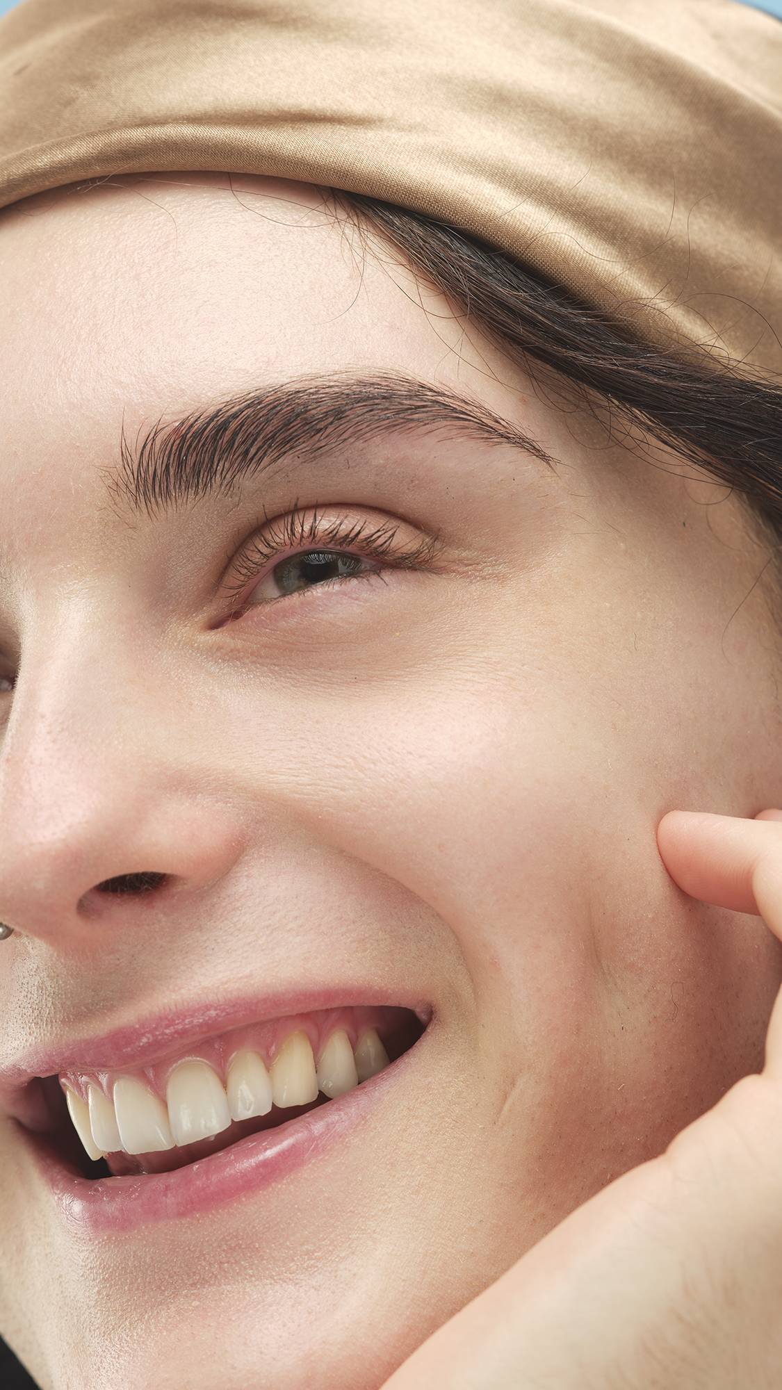 A close-up of a person with a satin gold headband, a nose ring, and well-defined brows. They are smiling and gently touching their cheek. Their eyelashes are coated with black mascara, likely Circus Lashes ~ Mascara, against a bright blue background.