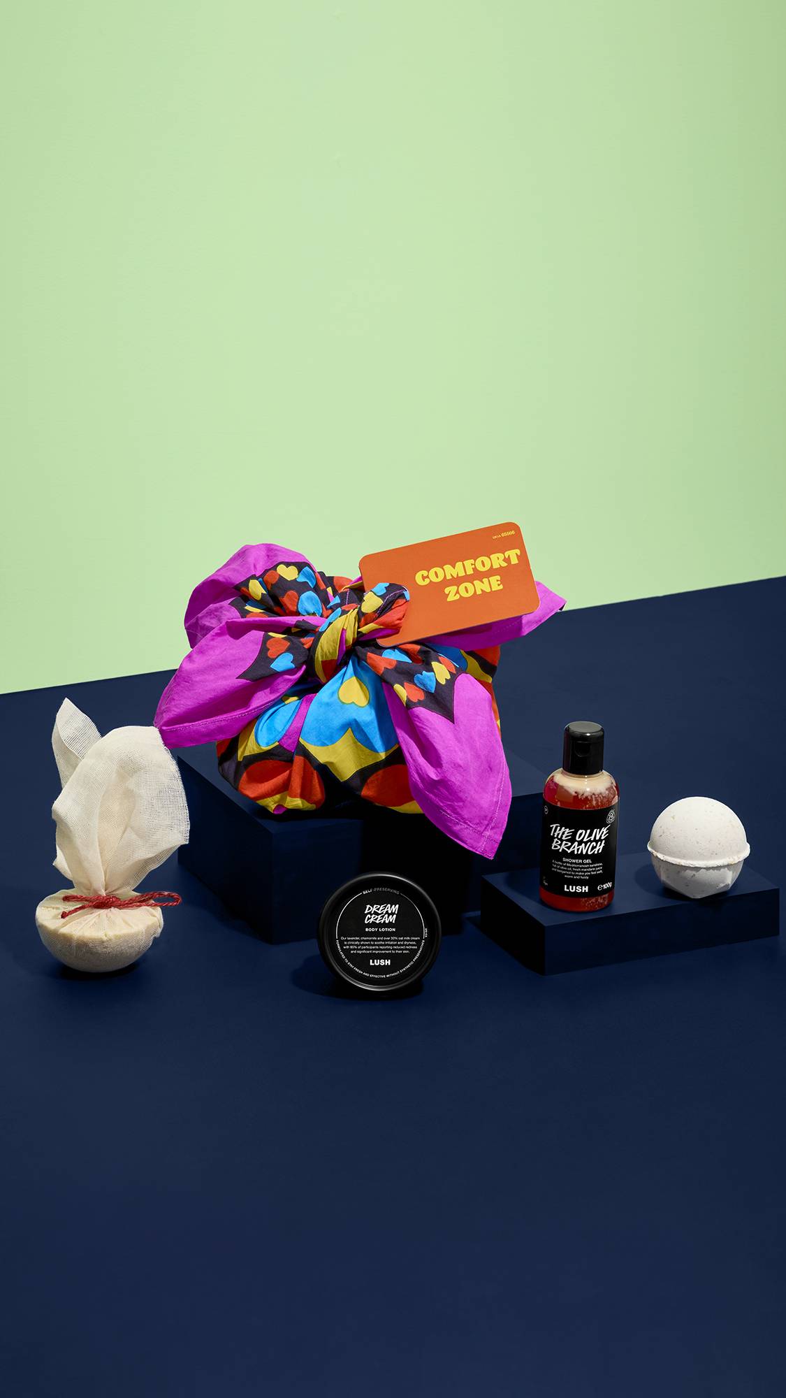 A knot-wrapped "COMFORT ZONE" gift set sits alongside unpacked products including Dream Cream, The Olive Branch shower gel, a bath bomb, and a wrapped bath melt, on dark blue pedestals against a pale green and blue backdrop.