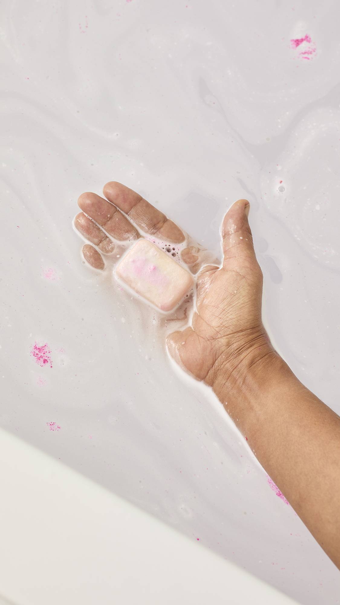 A top-down view shows a hand holding the pale pink "Creamy Candy" bath melt just under the surface of the water. The bathwater has transformed into a silky, milky white liquid with small pink distinct flecks floating around the dissolving bar.