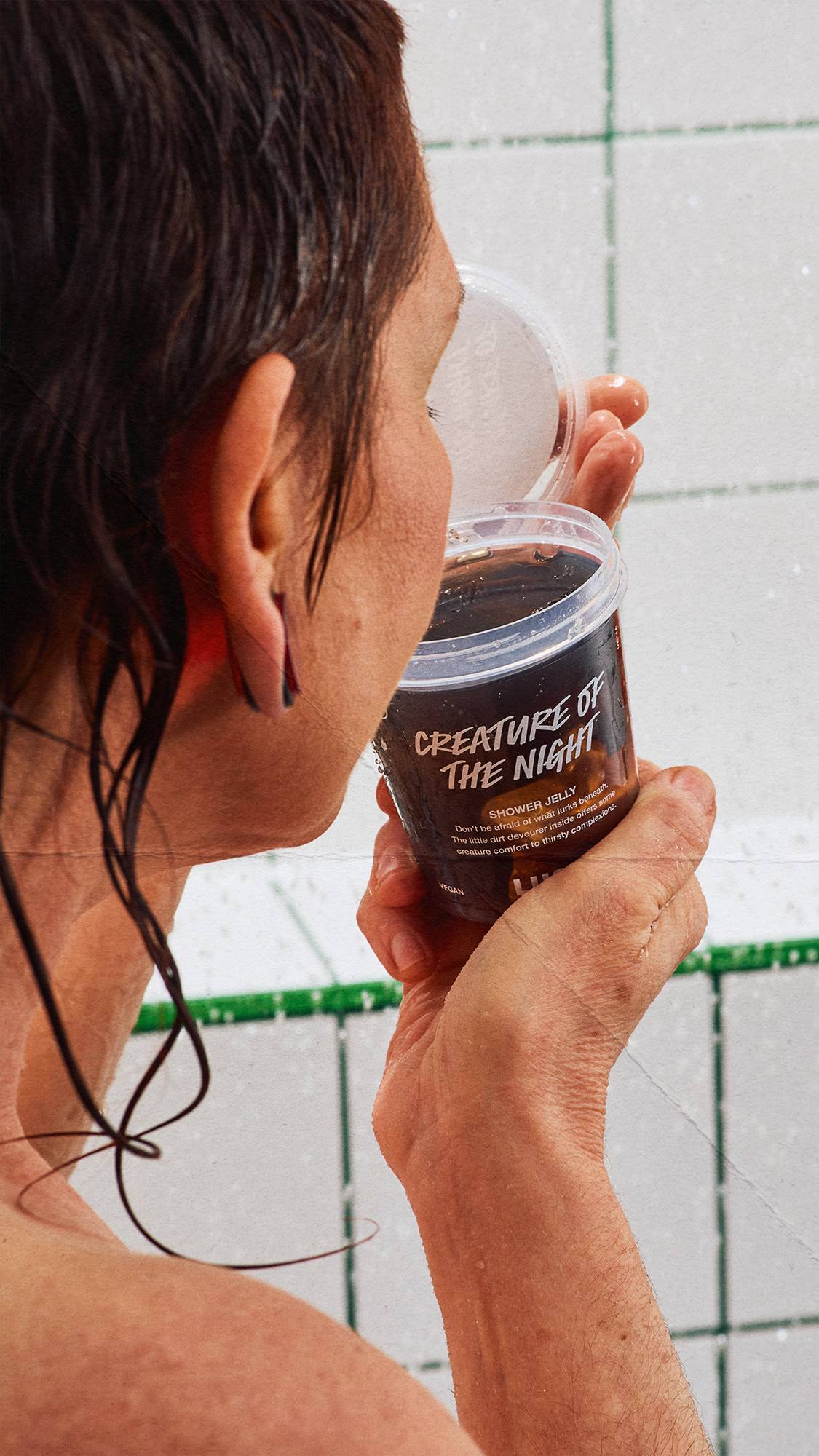 A side view of a person with dark, wet hair leaning in to smell an open tub of Lush 'CREATURE OF THE NIGHT' shower jelly in the shower. The background shows white square wall tiles with green grout lines.