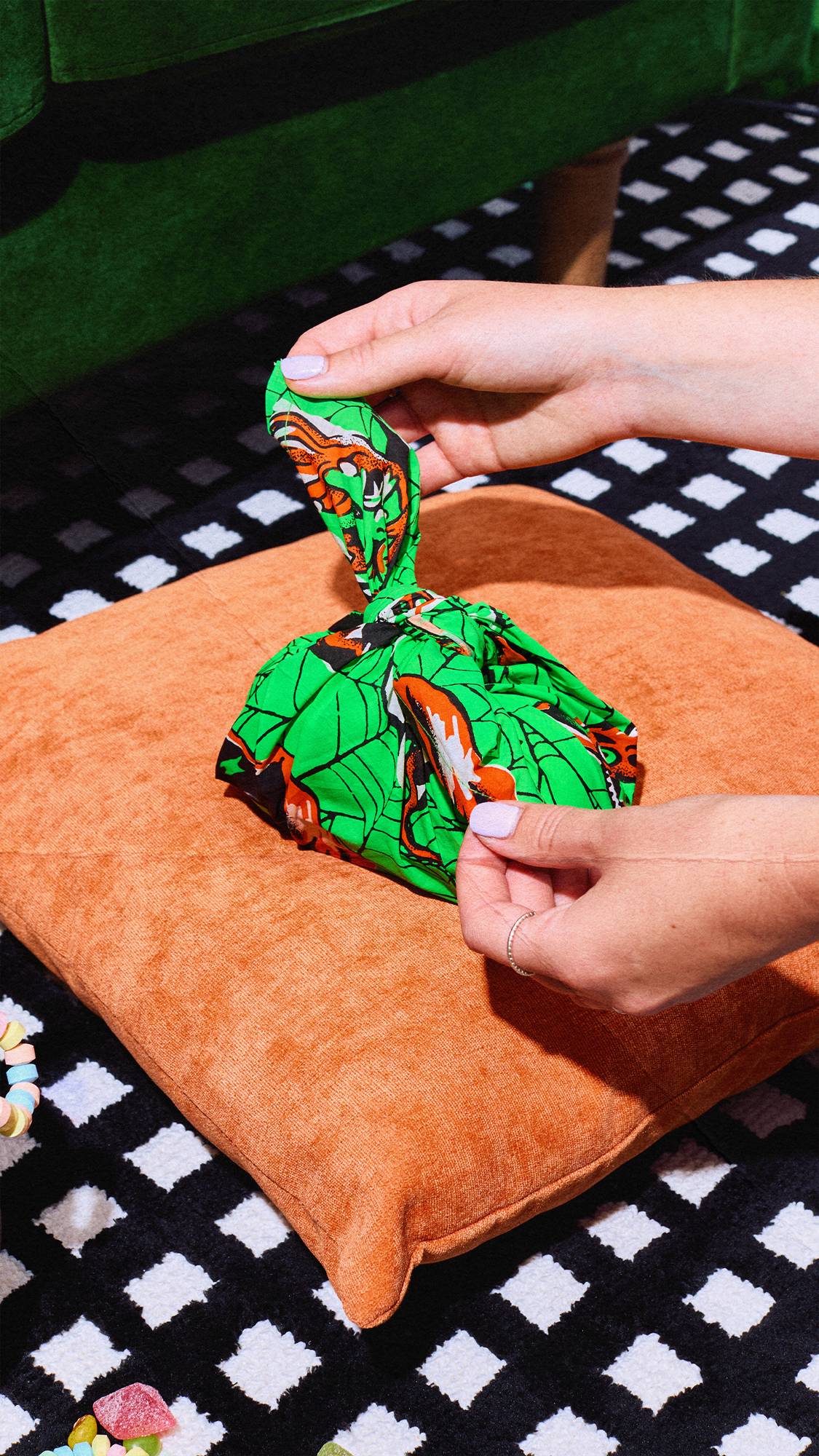 A person's hands untie a small gift wrapped in a vibrant green Halloween knot-wrap fabric. The gift rests on an orange cushion on a black-and-white chequered rug. Colourful sweets are scattered nearby, with a green couch at the top.
