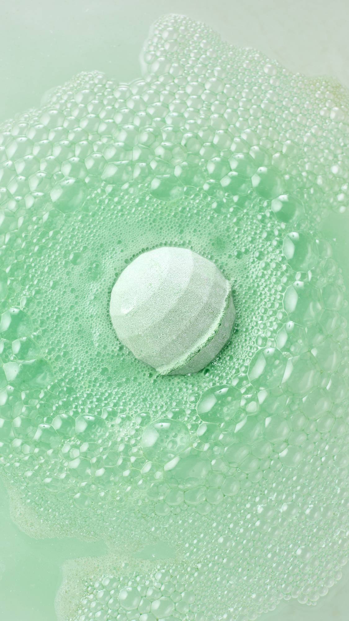 A round mint-green foot bomb floats at the centre of soft green water, generating a wide, expanding ring of pale green foam and frothy bubbles.