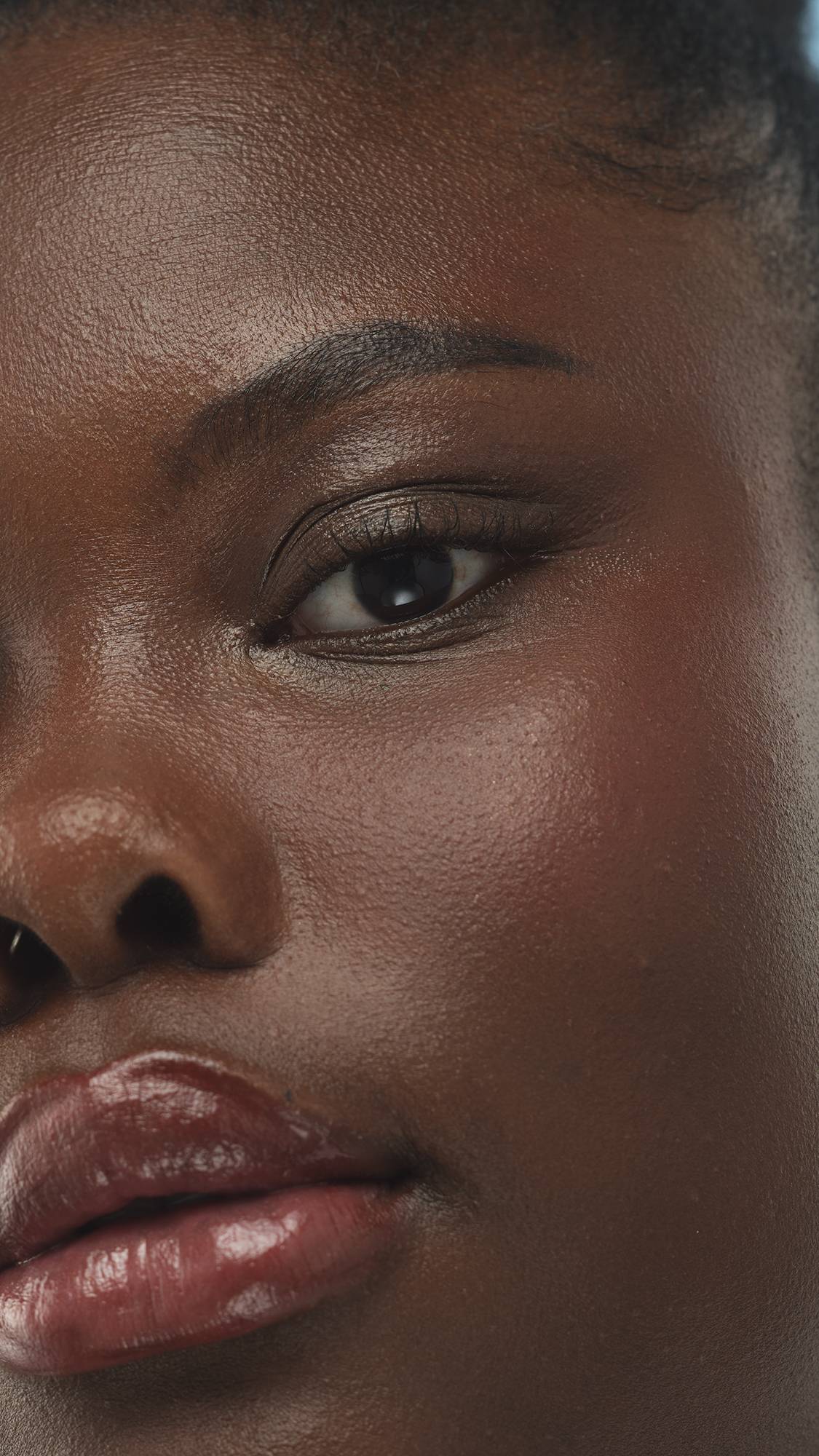 A close-up vertical shot of a person's eye, cheek, and lips, showing deep, cool-toned skin with a glossy finish from the Dark Cool Beauty Balm. The makeup enhances the natural contours of their face.