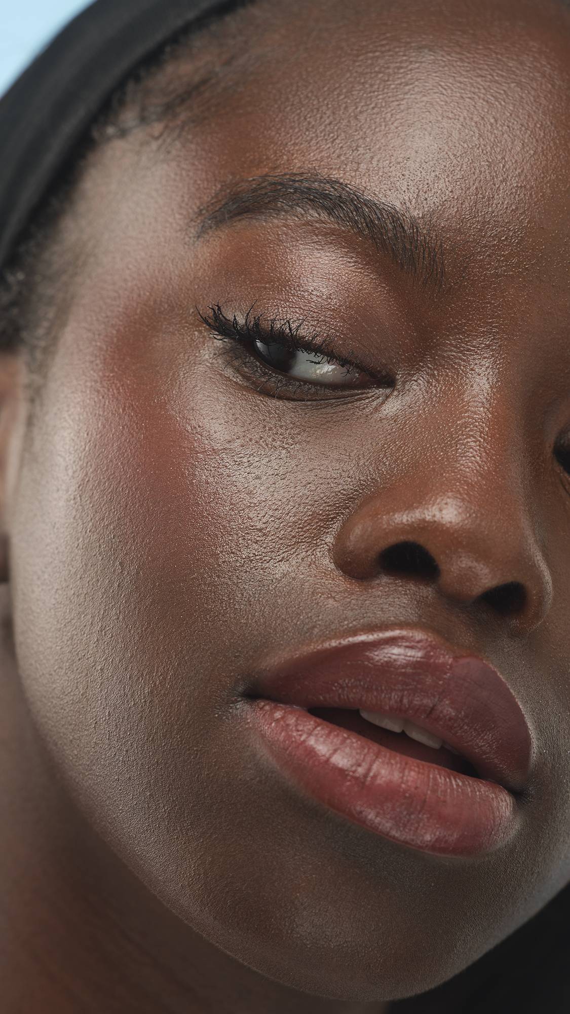 A close-up vertical shot of a person's upper face, focusing on their eye, cheek, and lips, showing a glossy finish and the Dark Neutral Beauty Balm product. They have deep skin and wear a black headband, against a soft blue background.