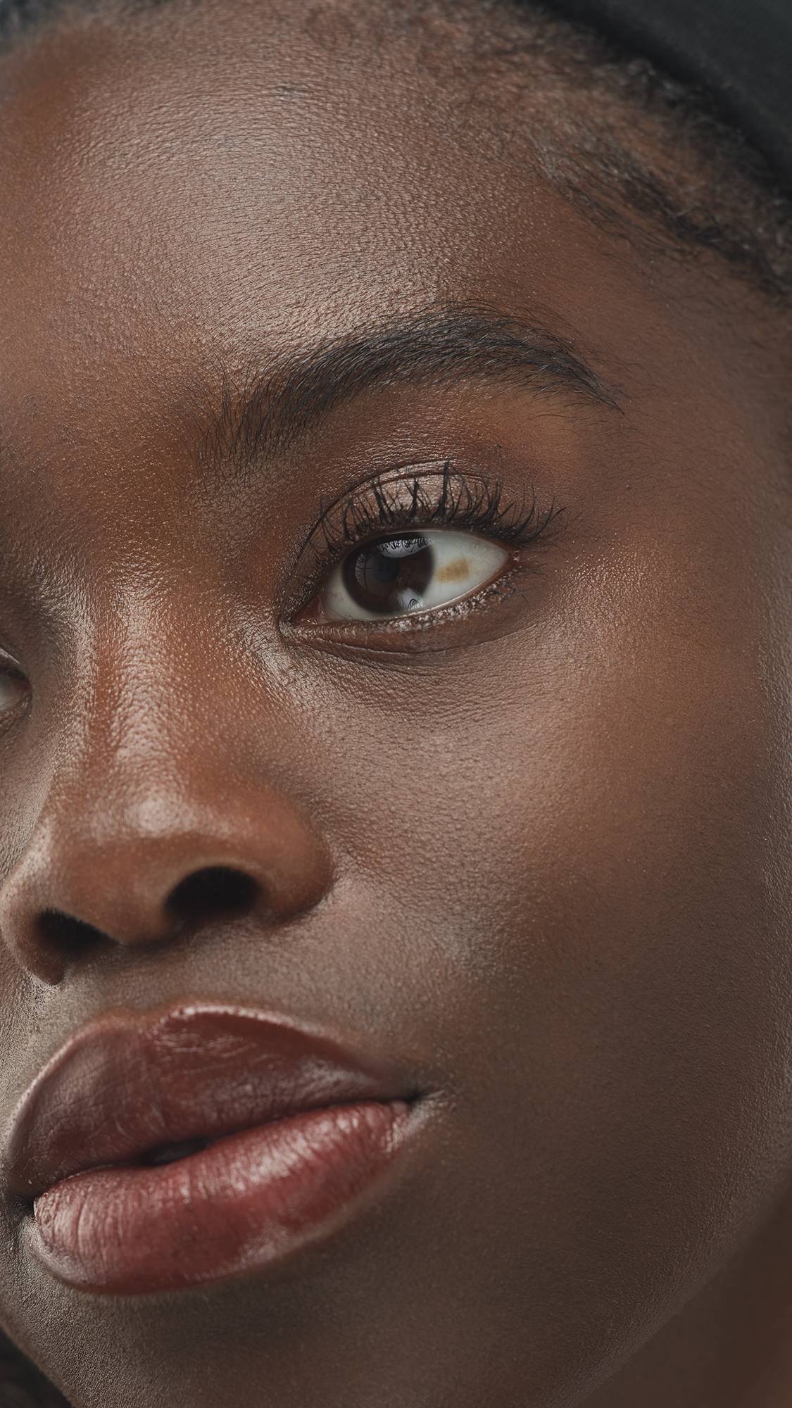 A close-up vertical shot of a person's eye and lips, showcasing a smooth, deeply-toned complexion with a subtle luminous finish from the Dark Neutral Skinwear product.