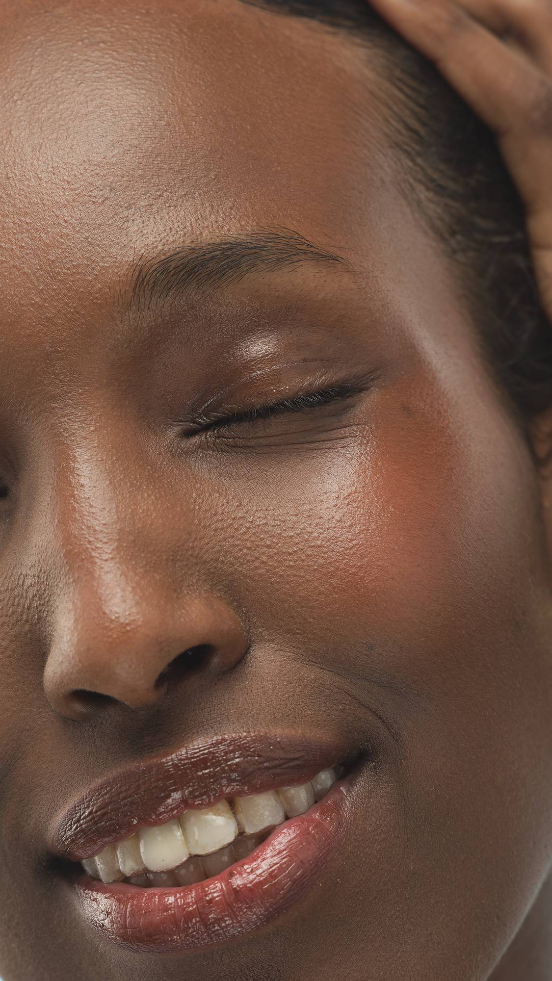 A close-up vertical shot of a person's smiling lower face, showing a glossy finish and the Dark Warm Beauty Balm product on their cheeks and lips. Their eye is closed, and they have deep, warm-toned skin.