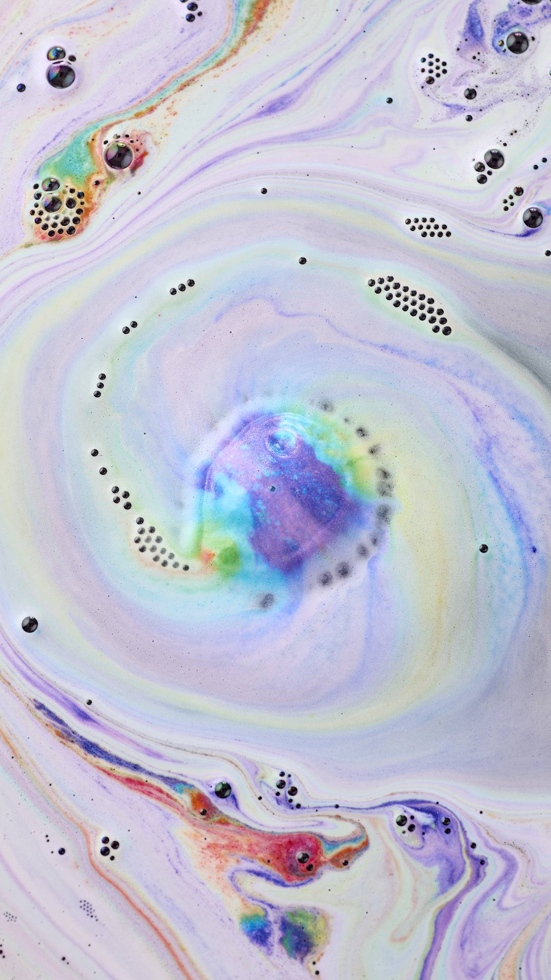 An aerial view of swirling, multi-coloured bath art created by the "Diversity" bath bomb. Spirals of purple, green foam create a marbled effect, with clusters of small bubbles and streaks of orange and blue dappled the surface.