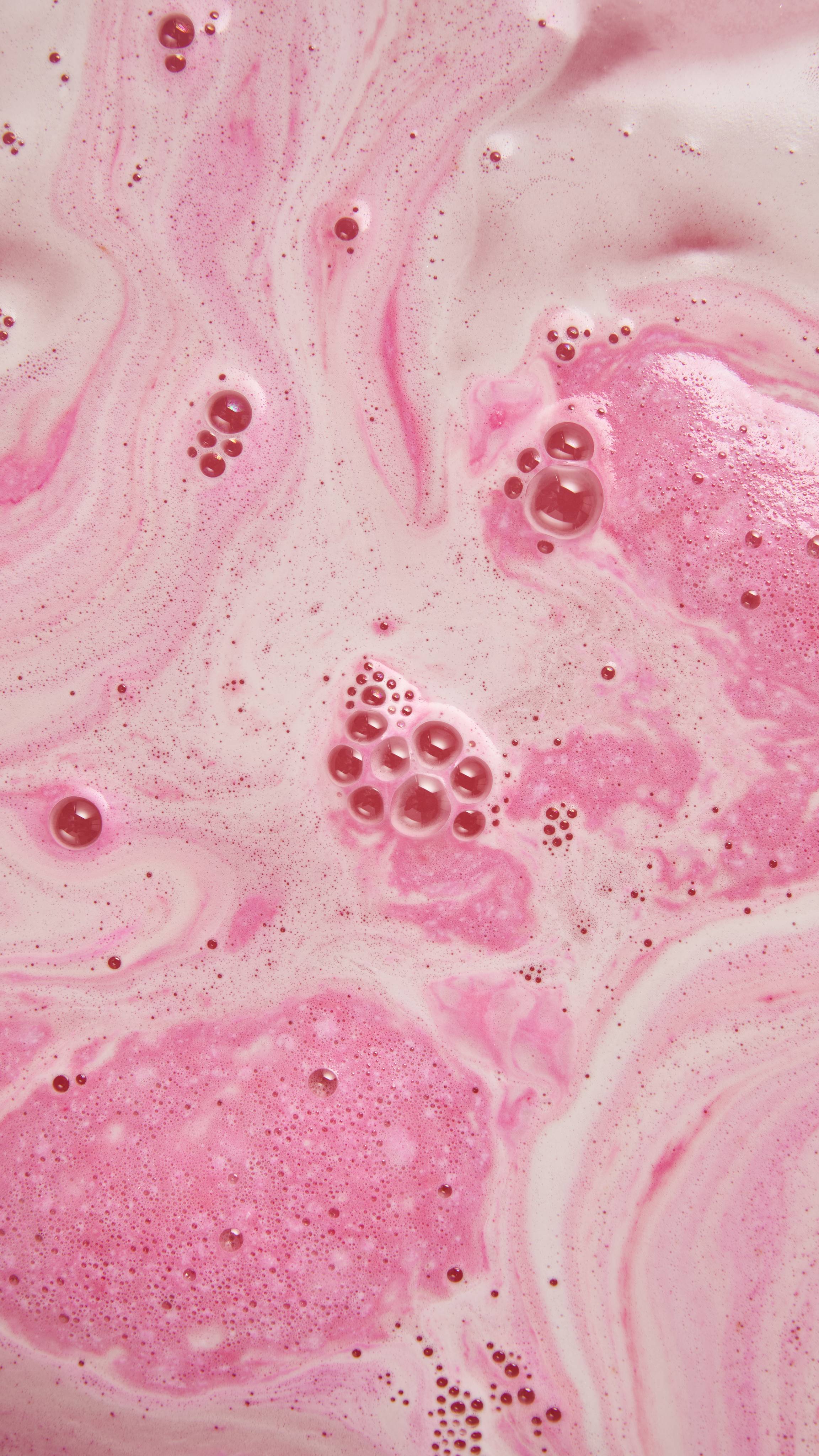 Swirling patterns of pale pastel pink and milky white bathwater, covered with patches of fine pink foam and small groups of glossy bubbles.