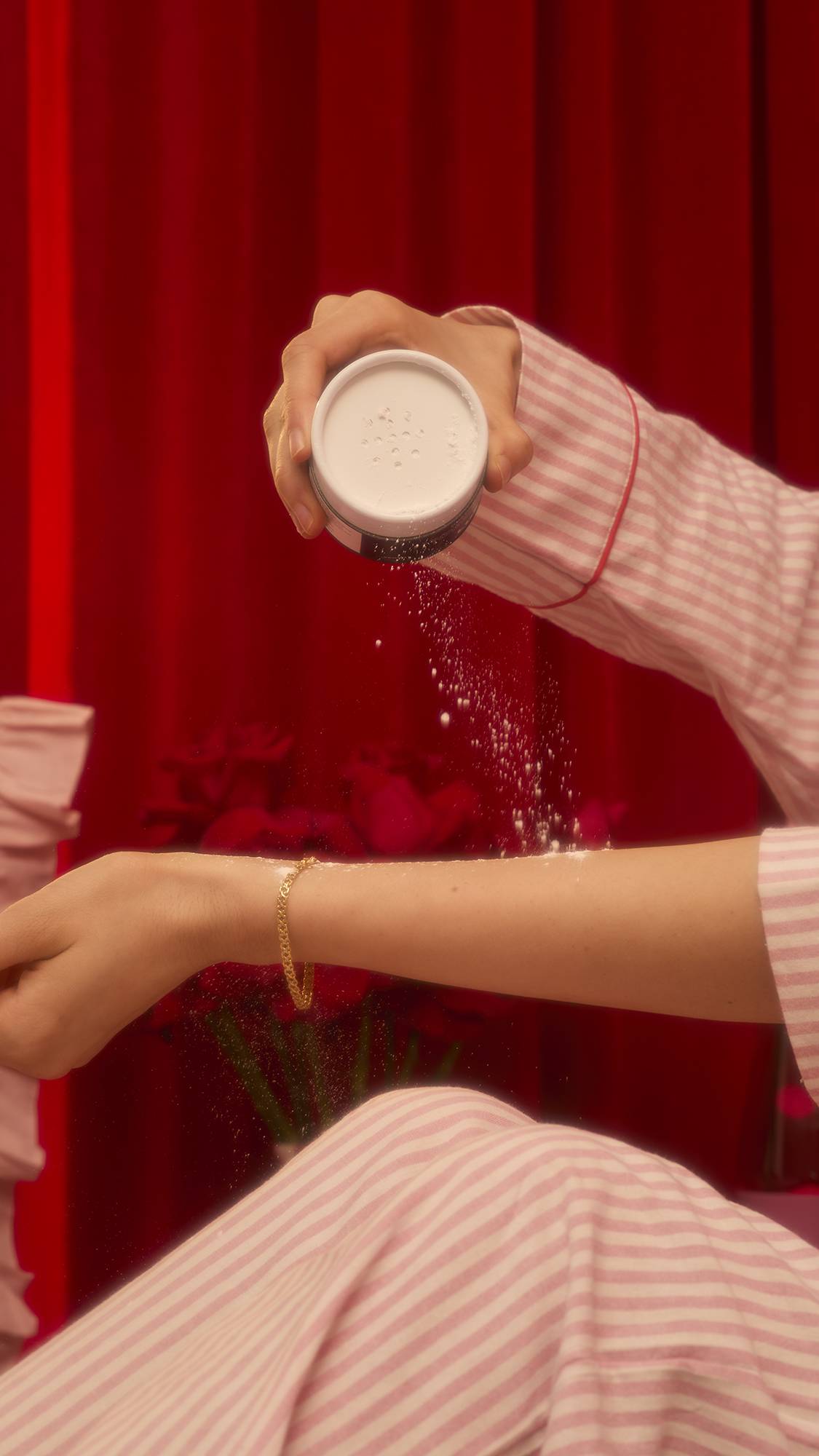 A person wearing pink and white striped pajamas shakes the white Dust The Two Of Us dusting powder onto their forearm, a gold bracelet visible on their wrist against a dark red curtain background.