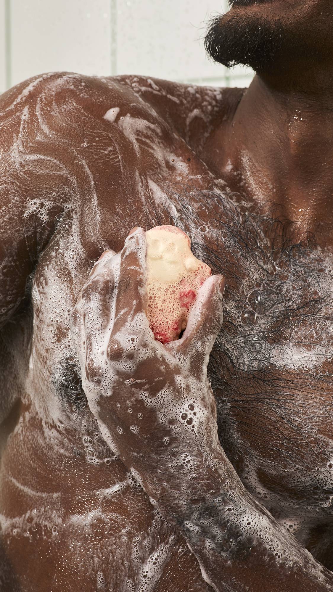 A model lathers a Santa Claus soap bar across their chest in the shower, covering wet skin with rich white bubbles against a white tiled background.