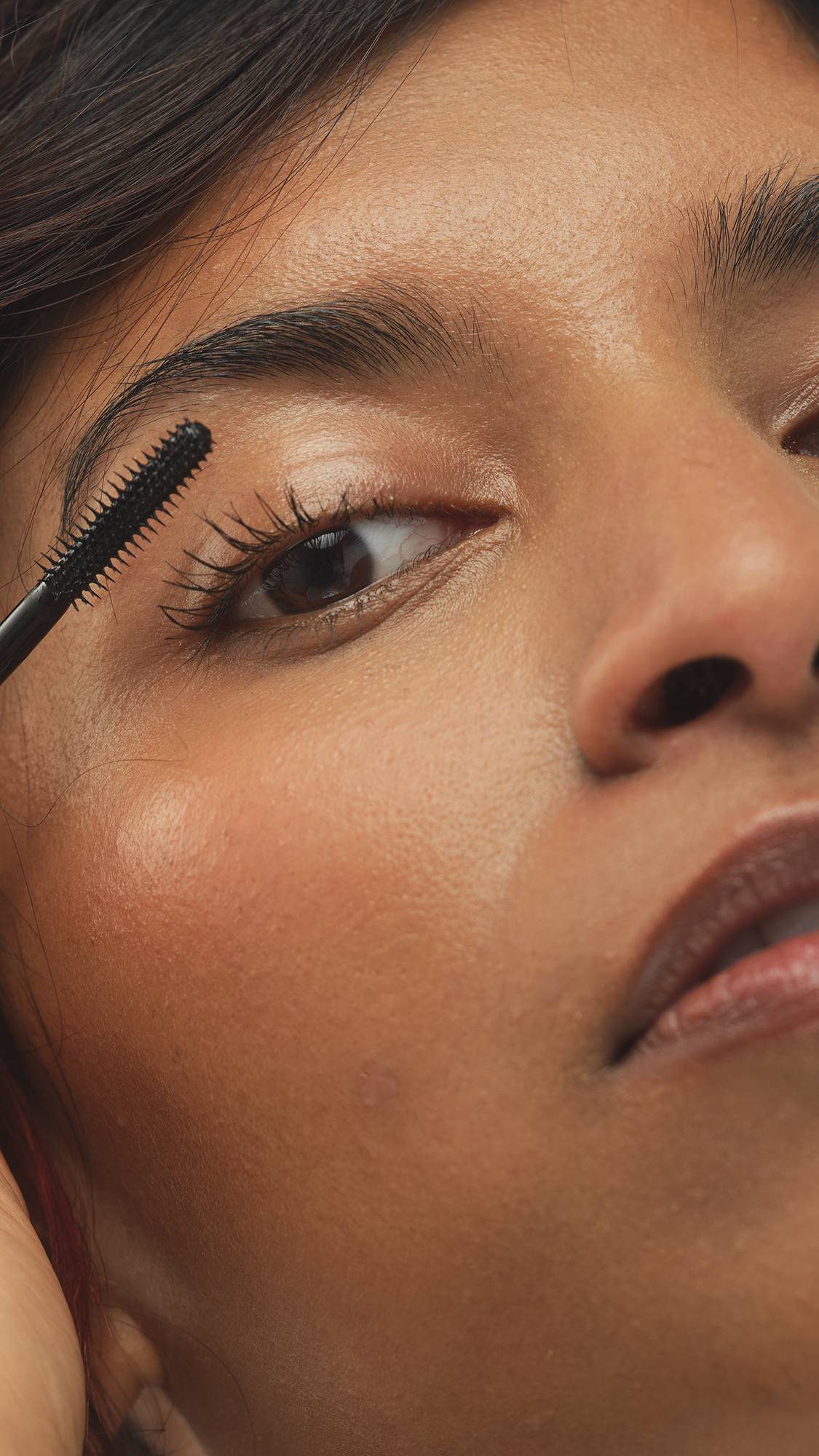 A close-up shot of a person applying Eyes Right ~ Mascara to their upper eyelashes with a black wand. They have defined eyebrows and a subtle peachy blush. The focus is on the mascara application.