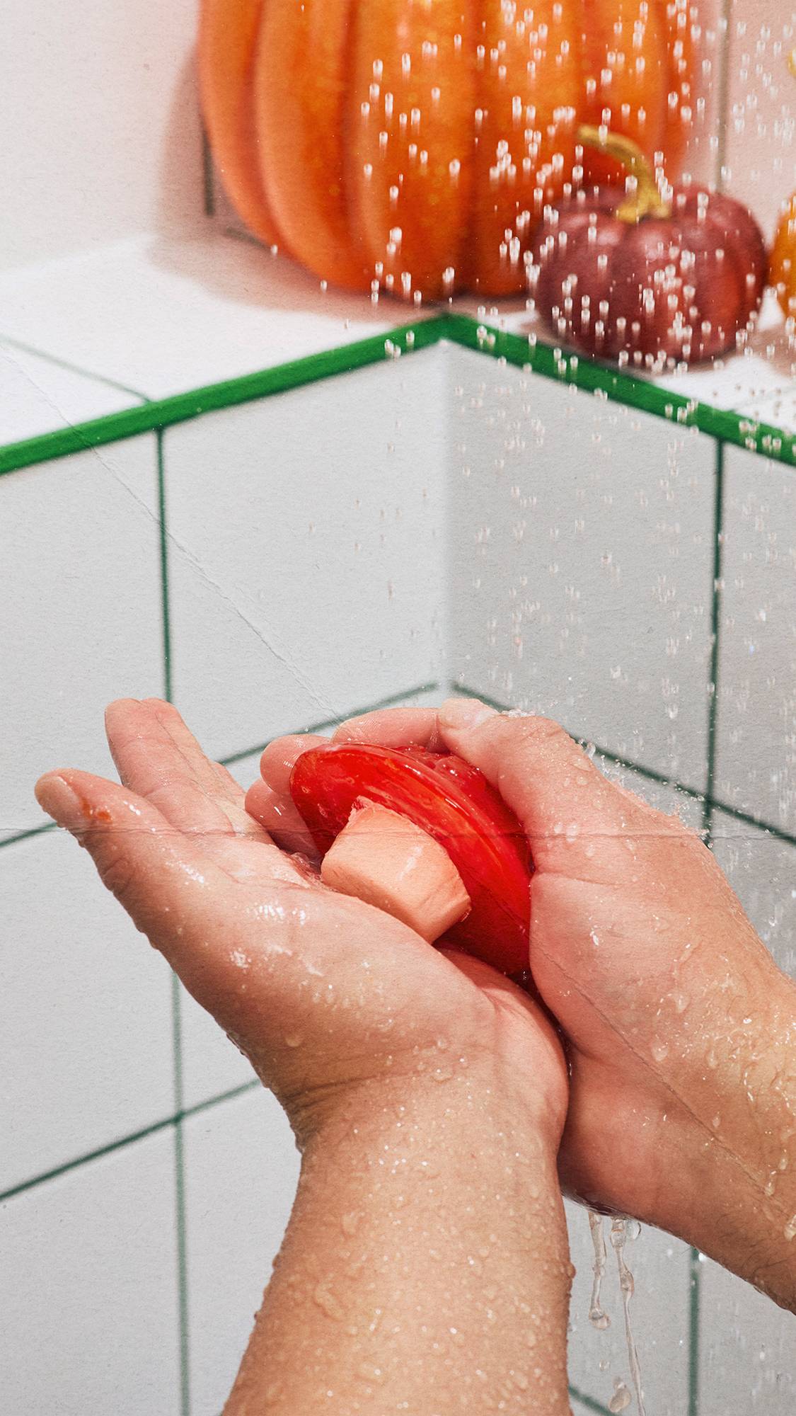 Water splashes over cupped hands holding a toadstool-shaped red soap in the shower, with green-grouted tiles and decorative pumpkins blurred in the background.
