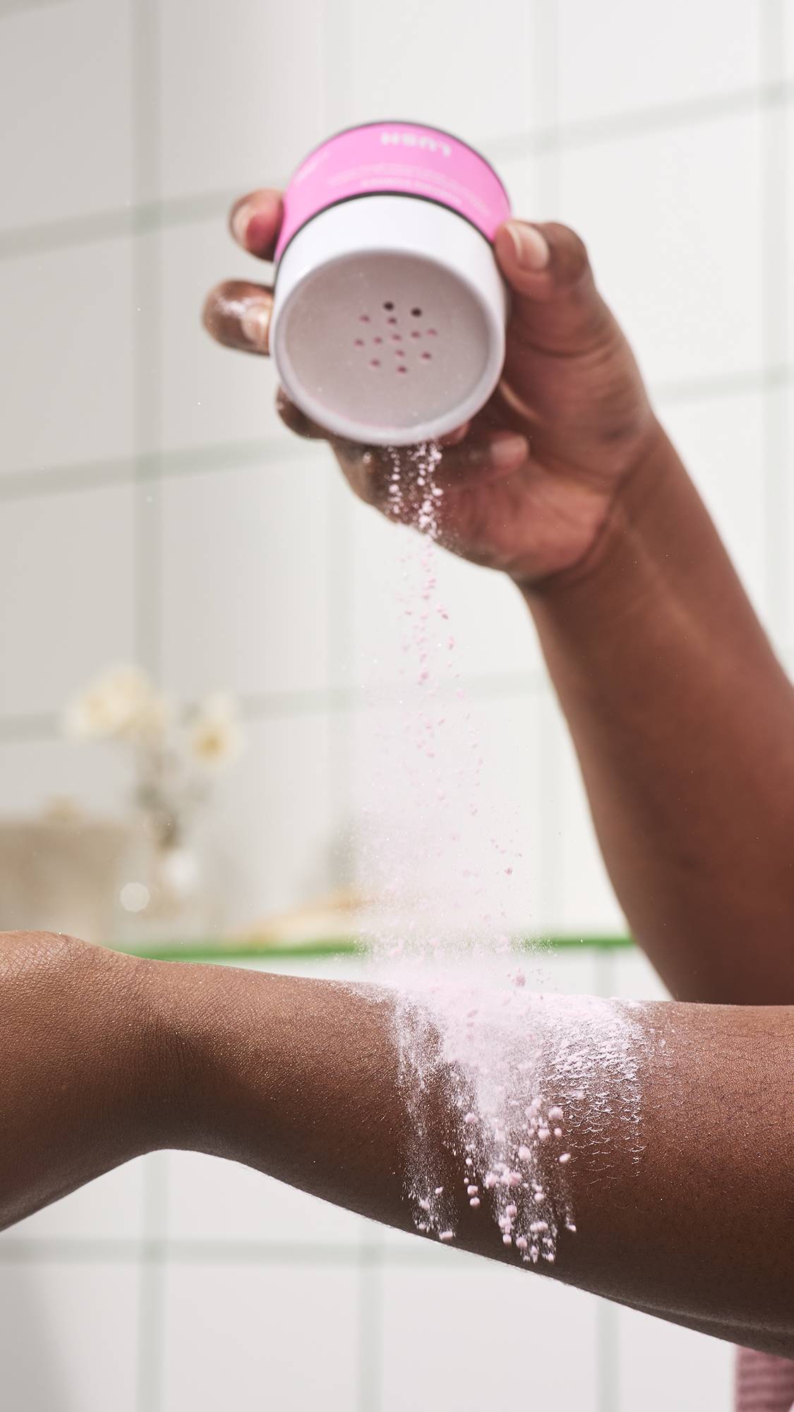 A hand shakes pink, Lush "Fairy Dust" dusting powder from a white tub with shaker holes onto an arm, leaving a light dusting of powder on dark skin against a white tiled background.