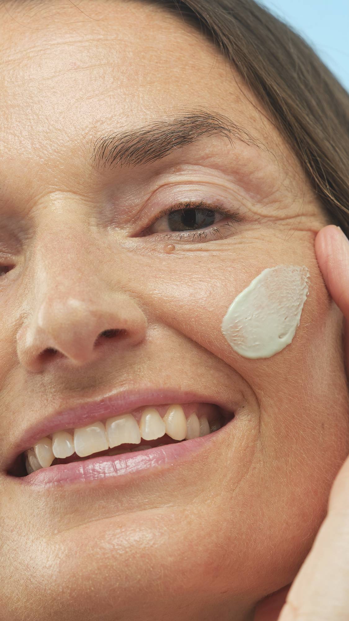 A close-up vertical shot of a smiling person's face, applying a creamy light-green swatch of Feeling Greener primer to their cheek with a finger. They have long brown hair and look slightly upward.
