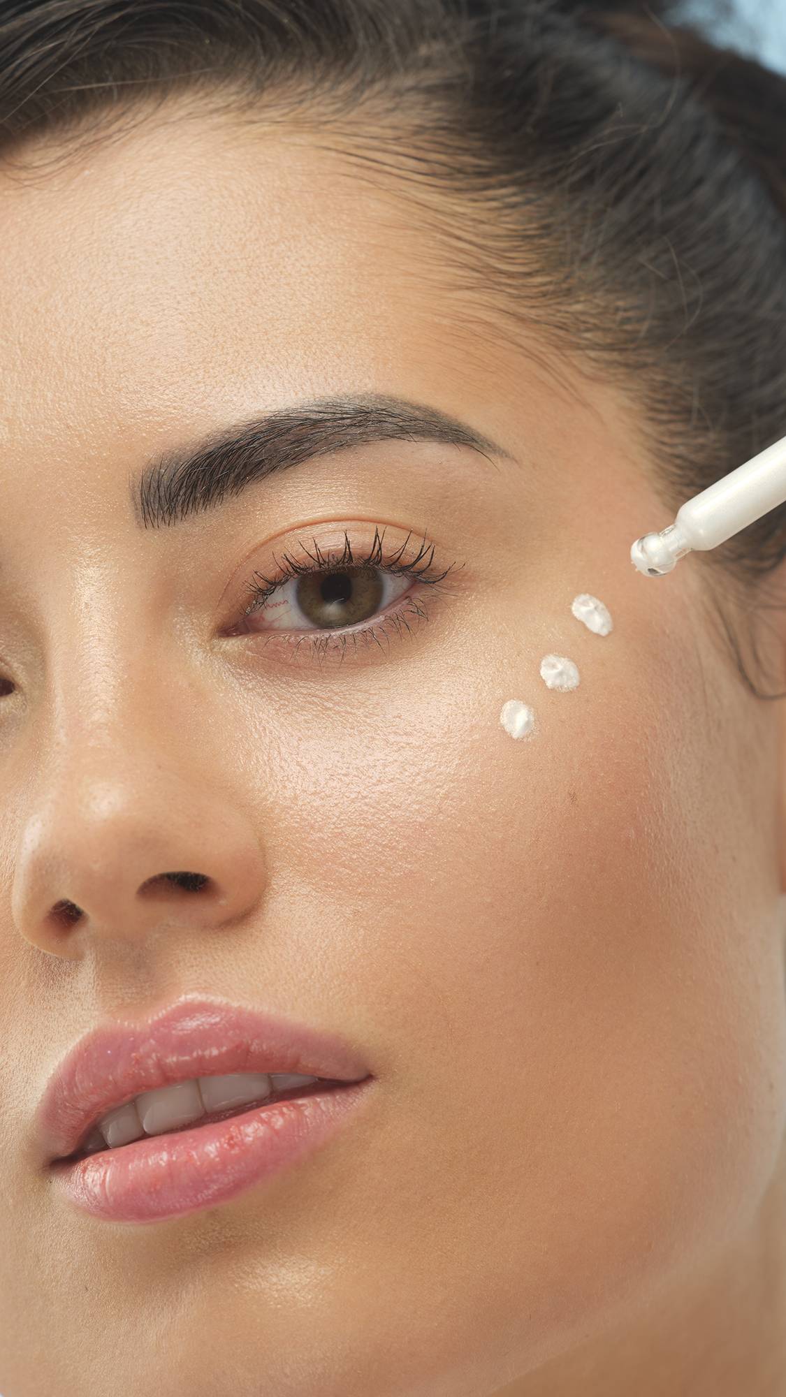 A close-up vertical shot of a person with medium skin, looking at the camera as three drops of the white Feeling Younger Skin tint are applied with a dropper to their cheekbone. The skin has a beautiful, dewy highlight.